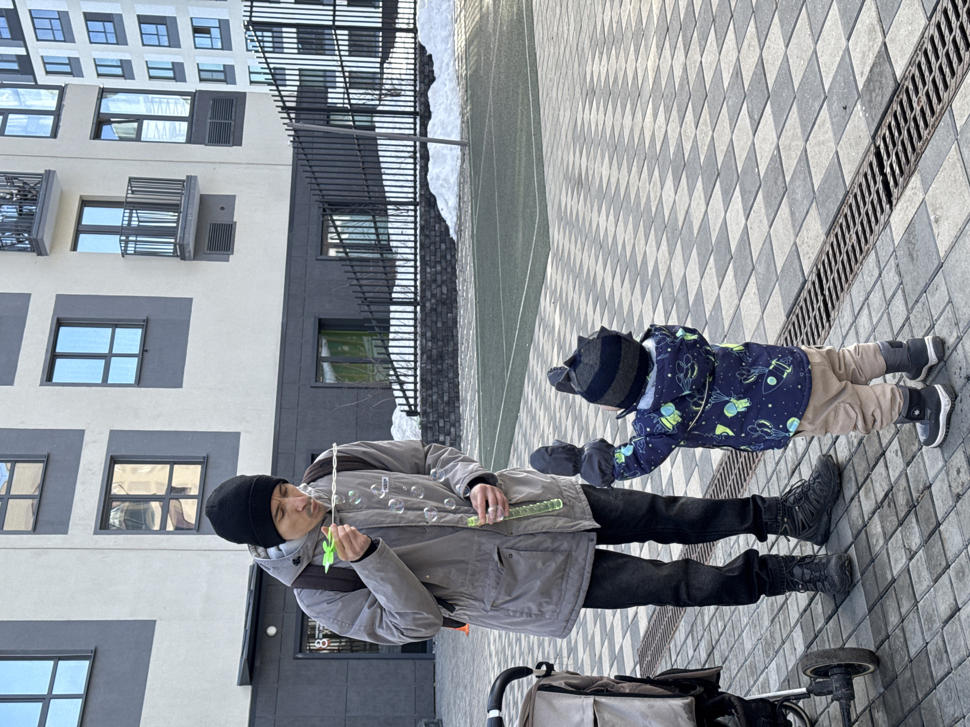 The image depicts an outdoor scene in a modern residential courtyard within Izhevsk, Russia. An adult male, wearing a black beanie, a grey hooded jacket, black jeans, and black boots, stands on the left. He has a dark backpack and is actively blowing iridescent bubbles from a green plastic wand and a transparent green bottle of solution. A young child, dressed in a blue winter jacket with a space pattern, tan pants, black winter boots, and a striped knit hat, faces the adult with arms raised, observing the ascending bubbles. To the adult's left, a beige baby stroller is partially visible. The ground is paved with a checkered pattern of light grey and dark grey interlocking pavers, featuring a metal drainage grate running horizontally. In the background, multi-story modern residential buildings with beige and dark grey facades and reflective windows are visible. A dark metal picket fence separates the paved area from a green-surfaced sports court with white lines and several piles of snow. The scene is illuminated by daylight, suggesting clear or partly cloudy weather.