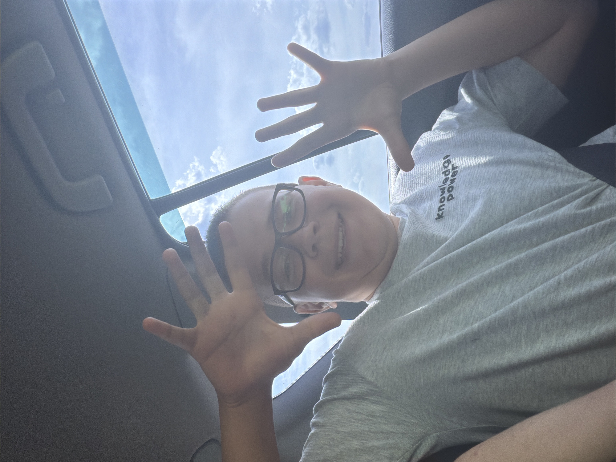 In a car, a young boy with short dark hair and glasses is taking a selfie. He is smiling and has his hands raised in front of his face. He is wearing a grey t-shirt with the words "Knowledge Power" printed on it. The boy is sitting in the back seat of the car, and the sky can be seen through the window. The scene is outdoors, and it appears to be a sunny day. The location is a car interior, likely on a road, with the sky and clouds visible through a car window. There is no indication of the city of Ostrovka, Russia, in the image.