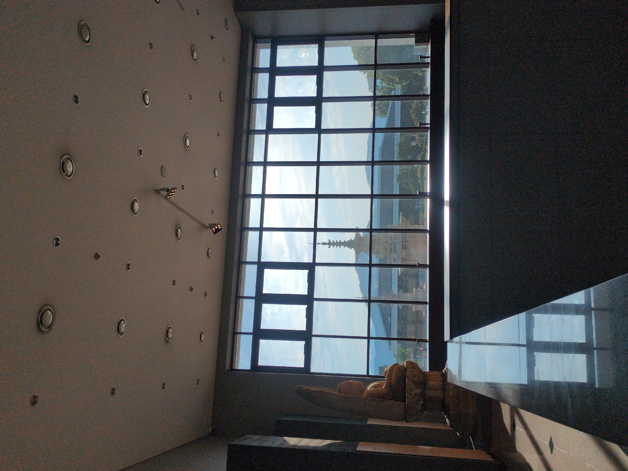 The image captures an interior scene within a building in Gyeongju-si, South Korea. A light gray wall on the left is adorned with numerous recessed circular lights. A long, adjustable arm fixture extends from the wall, ending in a pair of lights.
A large, multi-paned window dominates the central view, offering an exterior view. Through the window, one can see traditional Korean architecture, including a pagoda, set against a backdrop of distant mountains under a partly cloudy sky. The buildings suggest the area is historically significant, a characteristic feature of Gyeongju-si.
Below the window, a stone sculpture rests on a display platform. The figure's details are somewhat obscured by shadow.
To the right of the window is a dark wall and a large, reflective surface that mirrors the window and the exterior view. Light streams through the window, creating shadows on the floor. The overall ambiance suggests a museum or gallery setting.