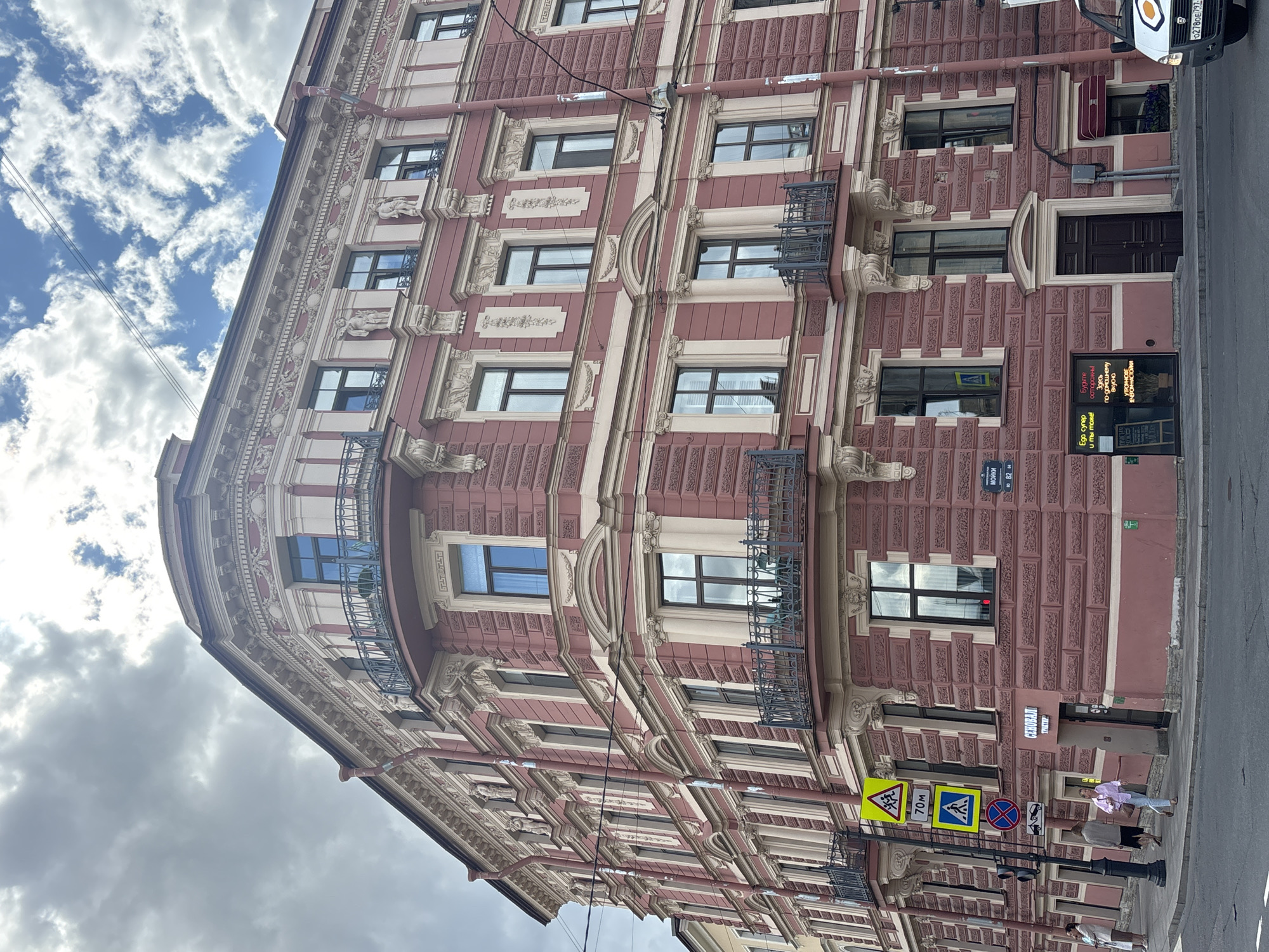 The image depicts a multi-story, ornate building on a street corner in Saint Petersburg, Russia. The building features a distinctive pink facade with cream-colored decorative elements, including pilasters, cornices, and sculpted figures above some windows. Several balconies with wrought-iron railings are visible. The sky is partly cloudy.
In the foreground, a street corner is visible with a pedestrian crossing and traffic signals. Several street signs are present, including a yellow diamond-shaped sign with children and an arrow, a blue rectangular sign with a pedestrian symbol, and a circular red and blue sign with a diagonal bar. A few people are walking on the sidewalk. One person in a pink top and light blue pants is walking away from the camera, carrying a handbag. Another person in a light-colored top and dark pants is walking behind them. A third person in a light-colored outfit is standing on the sidewalk to the left.
A storefront with illuminated text in Cyrillic script is visible at street level. The name "СЕНОВАЛ" is prominently displayed. A dark wooden door is located next to the storefront. A small awning with a dark red fabric is above a window to the right of the door. A white van with a partial license plate is parked on the street. The license plate appears to read "0278OE79".
The architectural style suggests a location in the historic center of Saint Petersburg, likely a commercial or residential building. The street signs and the Cyrillic text on the storefront confirm the location as Russia. The specific street is likely Moika Embankment, given the sign with Cyrillic text "МОЙКИ 82".