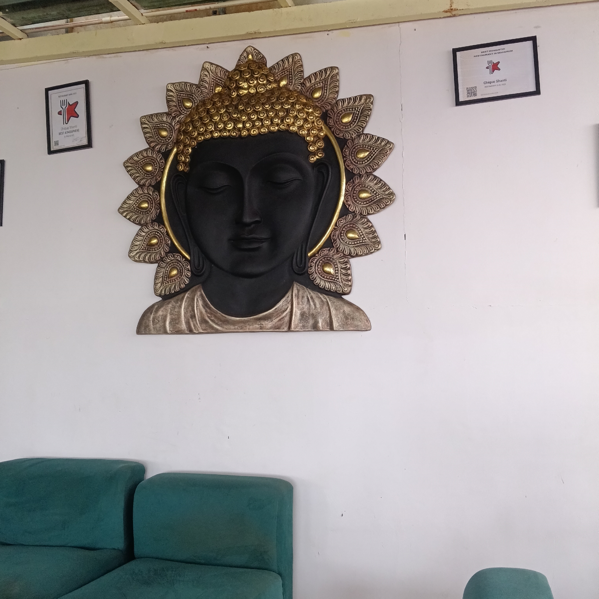 A wall with a Buddha head sculpture and two framed awards hangs above a green sofa, possibly in the city of Mandrem, India. The Buddha sculpture is predominantly black with gold accents. The frames contain text, and a logo with a fork and a star. The sofa is a deep teal green, and a portion of what appears to be an armchair or small sofa is visible on the lower right side. The wall is painted white.