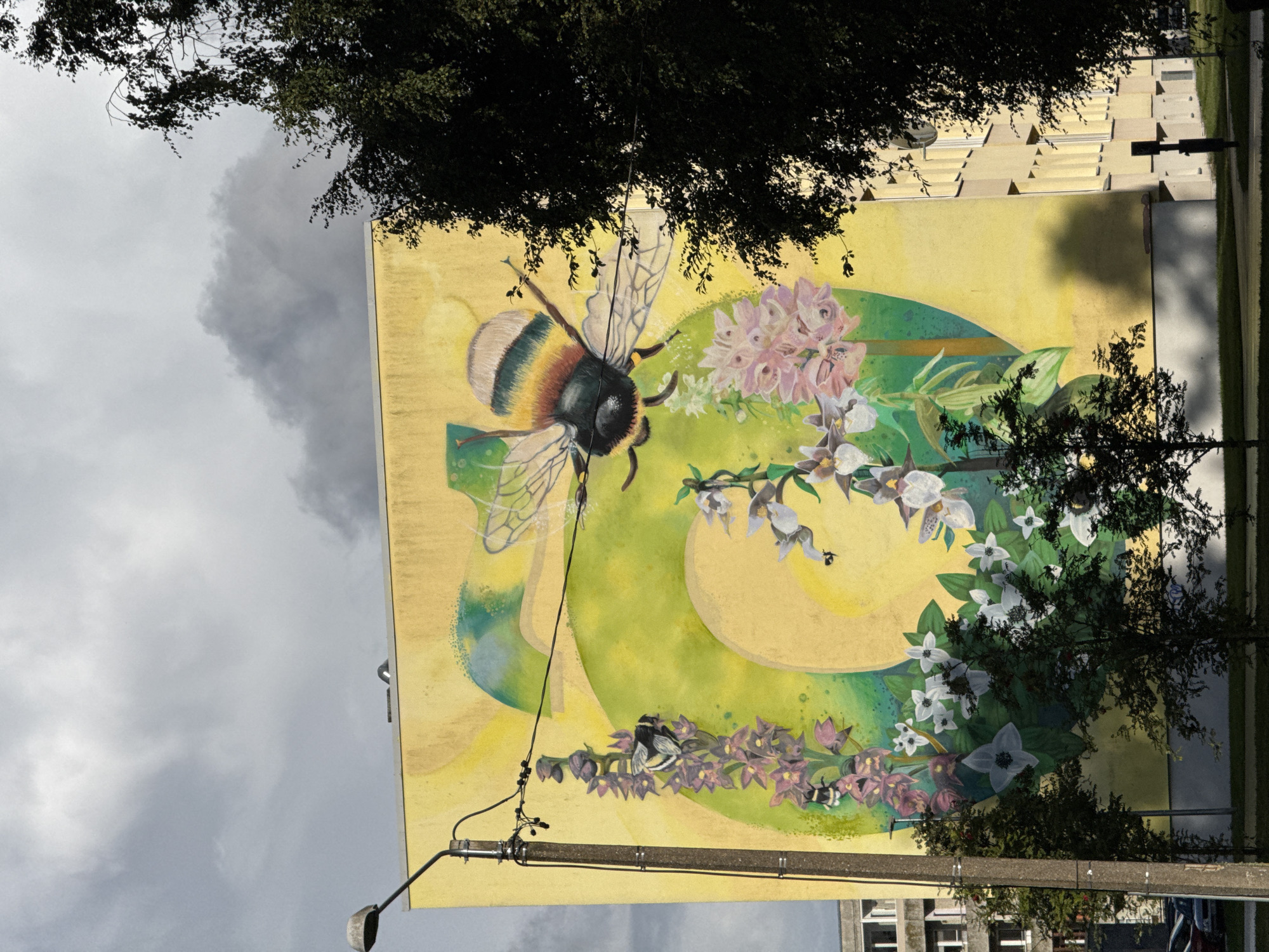 A mural on the side of a yellow apartment building depicts a large bumblebee with striped yellow, brown, and black colors, its wings spread, surrounded by stylized green and yellow shapes. Below the bumblebee, there is a cluster of pink flowers and several white flowers with dark centers. To the left, a tall purple flower stalk is visible with two smaller bumblebees on it. A lamppost stands in front of the left side of the mural, with black wires crossing the image. A dark green tree obscures the right side of the mural and the building. The sky above is overcast with dark grey clouds. The building is located in Tallinn, Estonia, as indicated by the architectural style and the presence of a typical apartment block building. The ground in front of the building is a grassy lawn, with a dark paved path or road in the foreground.