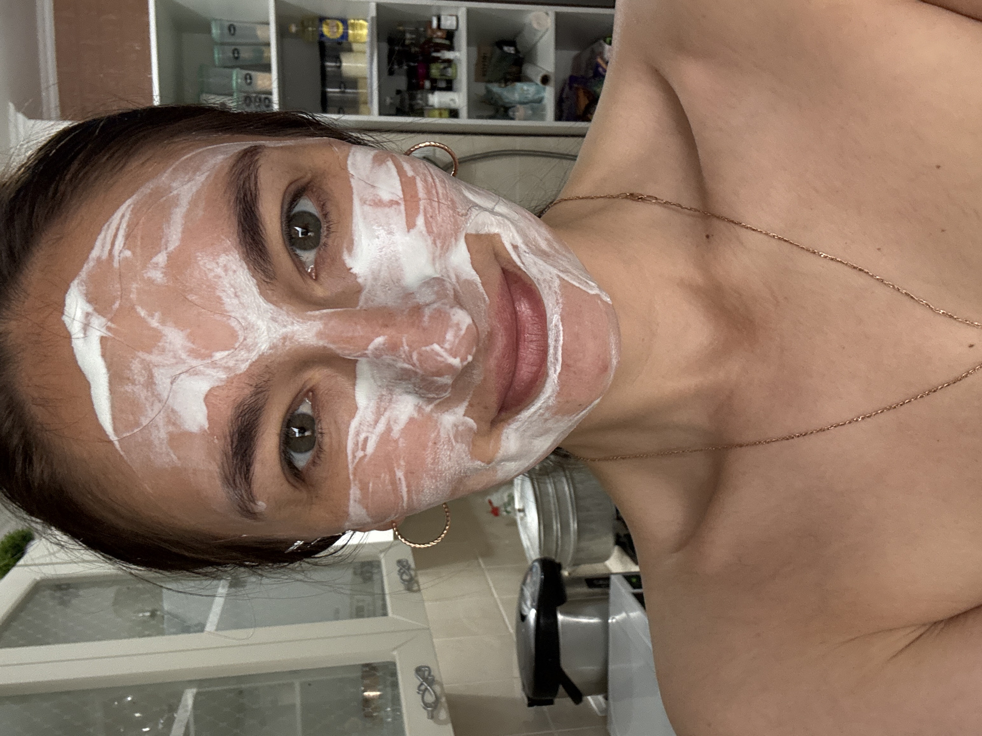 A selfie shows a woman with a white facial mask applied. She has brown hair, brown eyebrows, and olive green eyes. She is wearing gold hoop earrings and a delicate gold necklace. The background shows a kitchen interior with white cabinets and shelves containing various items. The presence of a kitchen appliance suggests an indoor domestic setting. The image provides no evidence of the city of Sochi, Russia.