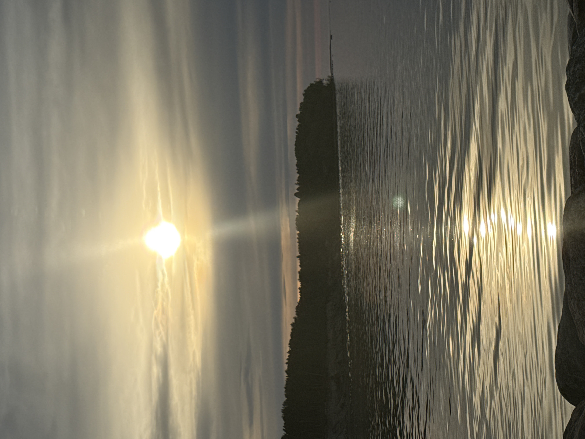 The image depicts a serene natural scene, likely a sunset over water, with a landmass covered in trees in the background. The sun is visible in the sky, casting a golden light that reflects off the rippling water. A rocky shoreline is in the foreground. The overall impression is one of tranquility and natural beauty. This scene is consistent with the coastal areas of Tallinn, Estonia, particularly locations like Pirita or the shores of the Baltic Sea. There are no people or specific objects identifiable that would allow for more precise identification within the city.