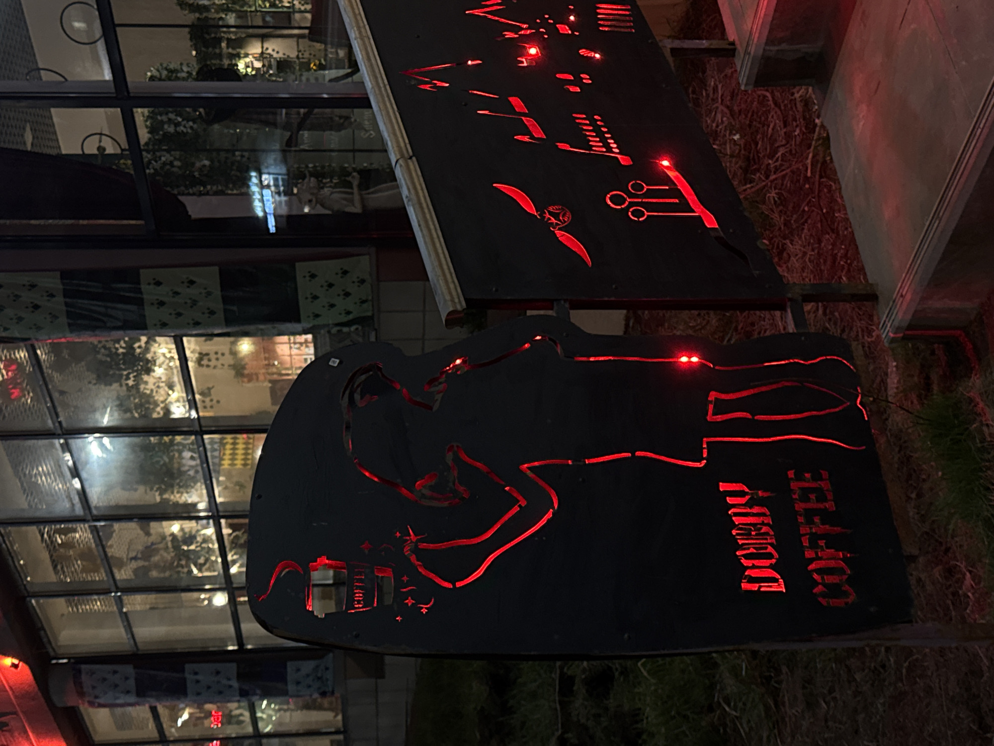 Here's a detailed description of the image:
**Setting:** The image depicts a nighttime scene outside what appears to be a café or coffee shop, potentially located in a city or urban area. The surroundings appear dark, with some vegetation visible.
**Objects:** Two black signs are prominently featured. These signs are lit with red lights, accentuating cut-out designs. The lower sign contains a figure, possibly holding a coffee cup, with the words "DOBBY COFFEE" vertically aligned. The upper sign has a more abstract design, resembling circuit board elements and a winged object.
**Environment:** The façade of a building, likely the café, can be seen in the background through windows. The reflections indicate a mix of natural light and artificial light sources within the building. The ground around the signs appears to be covered with grass or other ground cover.
