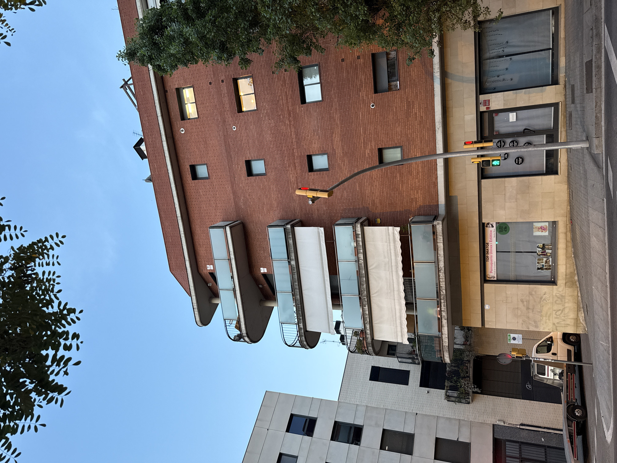 The image shows a street view of a residential building in Barcelona, Spain. The building is constructed with red brick and features several curved balconies with glass railings. In front of the building, there is a sidewalk and a street. A traffic light is visible, displaying a green light. A flatbed tow truck is parked on the side of the street. The building has commercial spaces on the ground floor, one of which appears to be a hair salon or beauty shop. The sky is clear and blue, with some tree branches visible at the top of the frame.