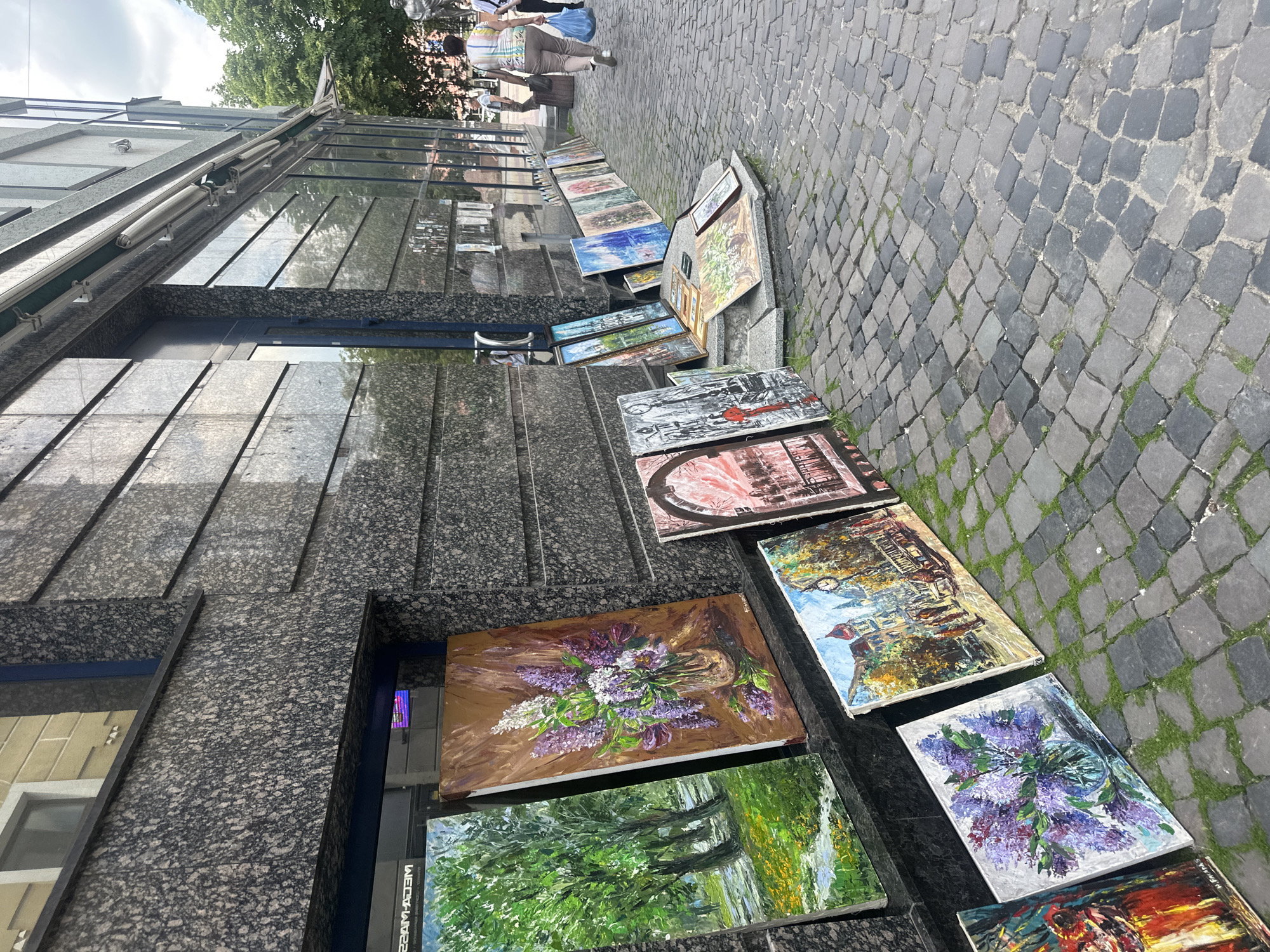 The image shows an outdoor art display with several paintings arranged along a cobblestone sidewalk next to a modern building with a granite facade. In the background, people are walking, and trees are visible, suggesting a public area. The paintings are diverse, featuring landscapes, still lifes (including a bouquet of lilacs and a colorful tree scene), and what appear to be cityscapes or abstract pieces. Some paintings are propped up, while others lie flat on the ground or on a low step. The overall scene suggests a street market or an artist selling their work. The time of day appears to be daytime, with natural light illuminating the scene. The weather seems to be overcast, indicated by the soft lighting and the sky's appearance. There are no discernible emotions conveyed by the people in the background, who are primarily focused on walking. No legible text is visible on the paintings or the building. The location context is Ternopil, Ukraine.