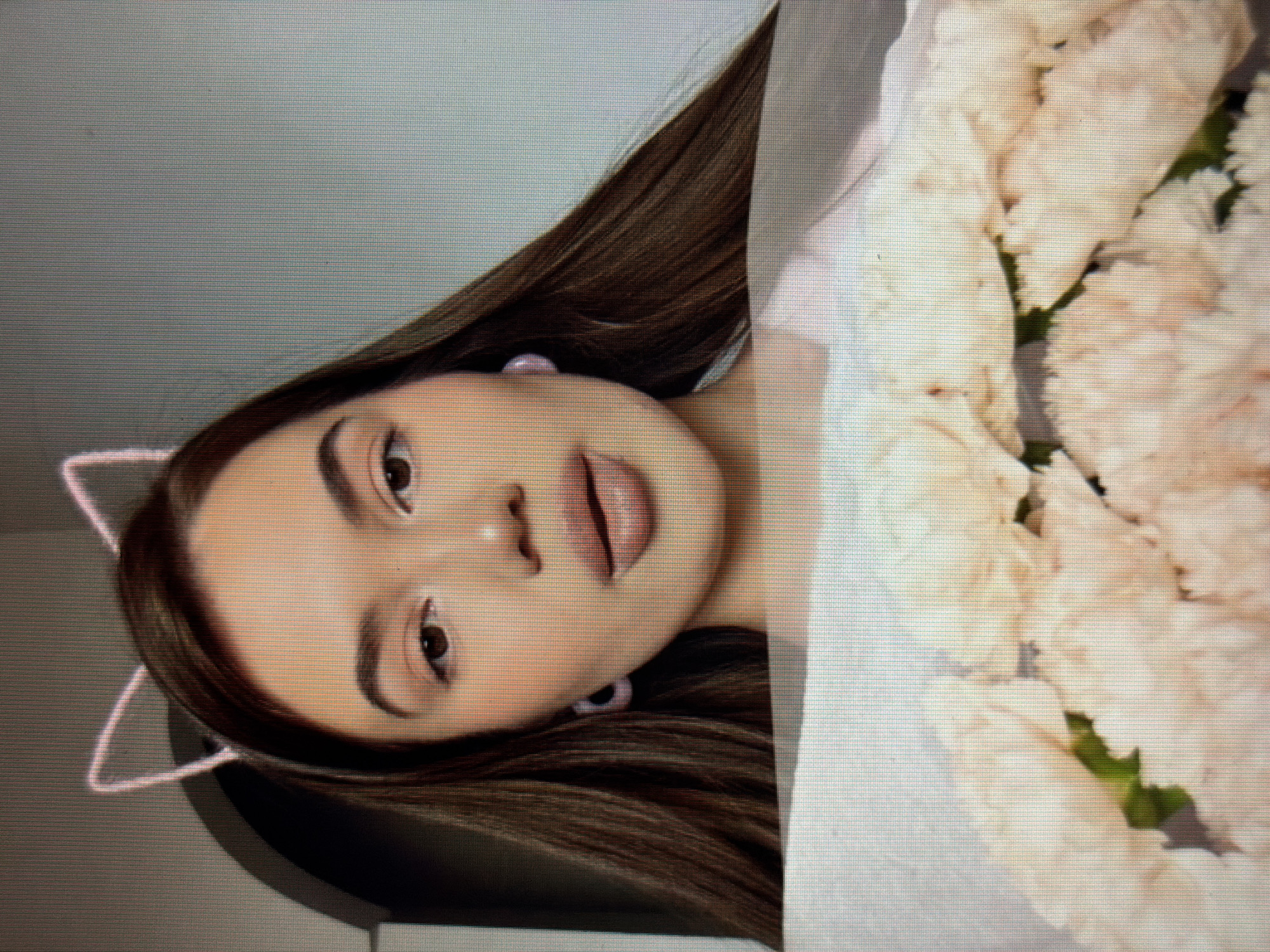 A medium shot shows a young woman with long, dark brown hair, wearing cat ear headphones. She has light skin, brown eyes, and is wearing subtle makeup. She has large, full lips painted in a nude shade and is wearing small, light pink, square earrings. She is holding a bouquet of pale pink flowers wrapped in white paper. The background is a plain, light grey wall. The image is pixelated, suggesting it was taken from a screen.