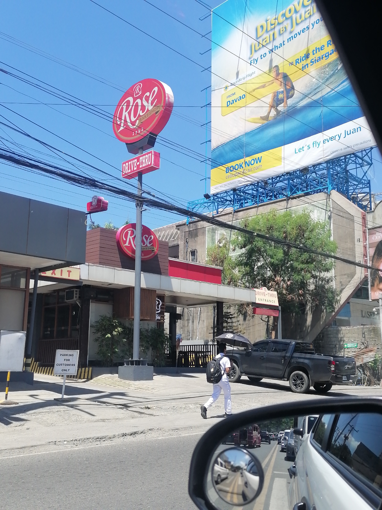 The image captures a street scene in Davao City, Philippines, likely near a commercial establishment identified by a prominent "Rose Bakeshop" sign with a "Drive-Thru" feature. The day is clear and sunny, with a bright blue sky visible above. Overhead, a network of power lines crisscrosses the frame.
In the foreground, a car's side mirror reflects a busy road with multiple vehicles in traffic. A white pickup truck is parked on the right side of the road. Further back, a person dressed in white pants and a white shirt, carrying a black backpack and holding a black umbrella, walks away from the viewer towards the bakeshop. The bakeshop itself has a red and white sign with the "Rose Bakeshop" logo and a "Drive-Thru" sign above it. An "EXIT" sign is also visible on the building. To the left of the bakeshop, a sign reads "PARKING FOR CUSTOMERS ONLY".
A large billboard advertising "Discover Juan" is prominently displayed in the background, promoting flights from Davao. The billboard features an image of a surfer. The overall environment suggests an urban setting with commercial activity and traffic.