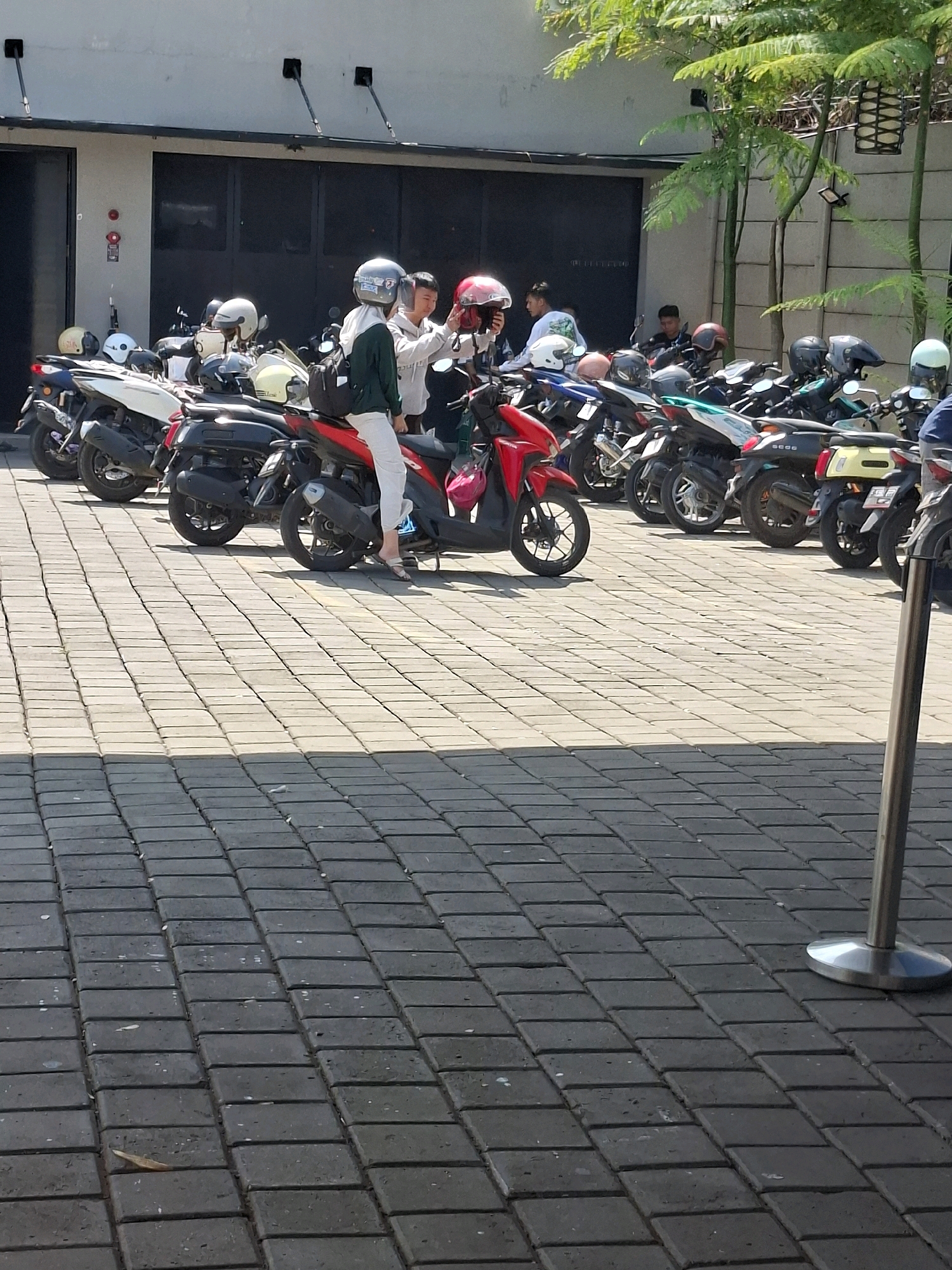 In Cianjur, Indonesia, a paved outdoor area, possibly a parking lot, is filled with numerous parked motorcycles. The scene is under bright daylight, casting distinct shadows on the brick-patterned ground. In the foreground, a person wearing white pants and a green jacket is seated on a red scooter. To their left, another person in a gray jacket is standing next to a white and black scooter. A cluster of motorcycles extends into the background, with some individuals standing around them. The environment suggests a public space, possibly near a commercial establishment or event venue, given the concentration of motorcycles and people. The architecture in the background includes a light-colored wall with dark doors or windows, and greenery is visible above.