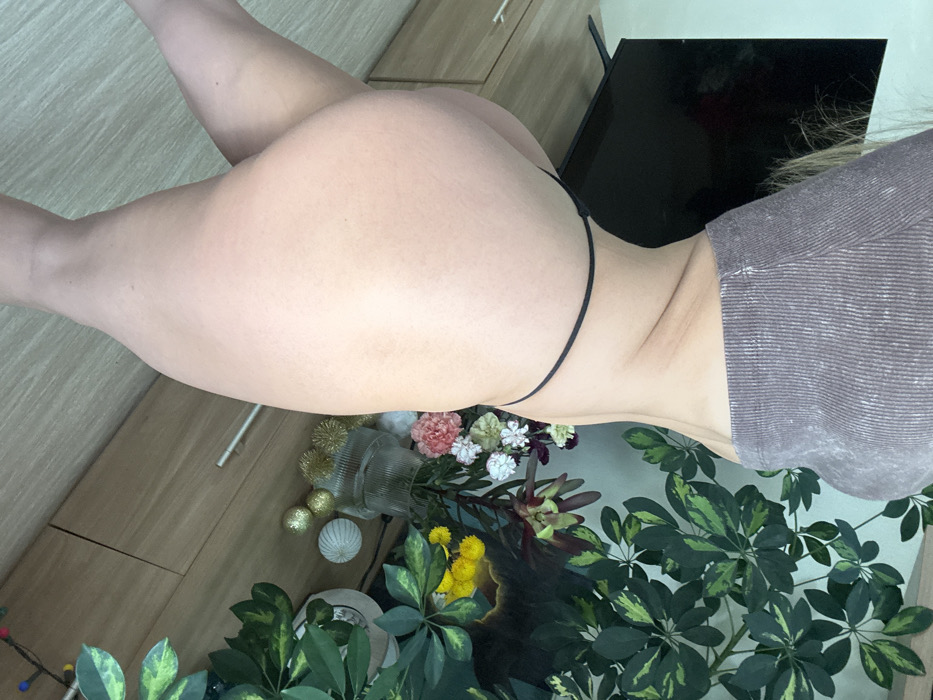 The image captures an indoor scene depicting a single individual, identified as an adult female. The subject is viewed from a slightly elevated perspective, showcasing her back and left side from mid-torso to mid-thigh. She is wearing a ribbed, short-sleeved cropped top in a muted grey-brown color and a black g-string. Her body is positioned with her back partially turned towards the camera, suggesting a pose.
The background features a contemporary residential interior. A dark flat-screen television on a stand is visible on the left, resting on a light-colored wooden media console. This console has at least three distinct storage compartments with horizontal handles. To the right of the console, a clear, fluted glass vase contains a floral arrangement comprising white, pink, and dark red blossoms, with additional yellow flowers partially visible behind it. Three golden, spherical decorative ornaments with a spiky texture and one white, textured spherical ornament are arranged on the console surface. A large houseplant with variegated green and yellow-green leaves occupies the upper right portion of the frame. The flooring is a light-colored wood or laminate, and the wall behind the console is a plain, light tone. The scene is illuminated by ambient, uniform lighting.
No visual identifiers within the image allow for the forensic confirmation of its specific geographic location as Saint Petersburg, Russia.