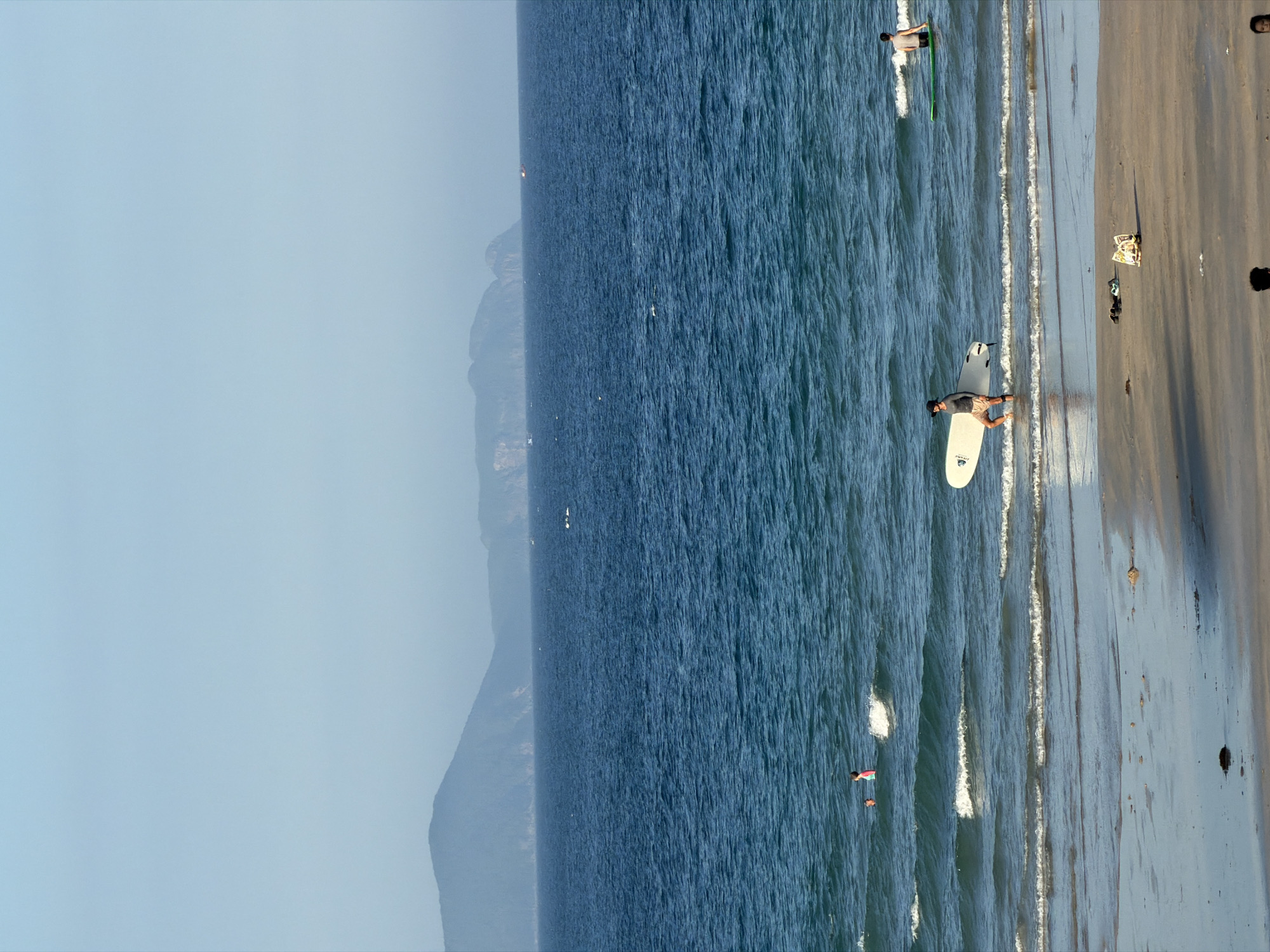 The image depicts a coastal scene in Cam Lam, Vietnam, likely on a bright, clear day. The foreground is a sandy beach with a few scattered objects, possibly debris or personal belongings. The water is a vibrant blue, with gentle waves lapping at the shore.
In the water, several individuals are visible. To the right, a person is standing in the water with a surfboard. Further to the left, a person is walking out of the water carrying a surfboard. Two other people are in the shallow water to the left, one appearing to be a child in a pink swimsuit.
In the distance, the expansive blue ocean stretches to the horizon, where a hazy line of mountains forms the backdrop. The sky is a pale, clear blue, with no clouds visible. The overall impression is one of a serene beach day.
