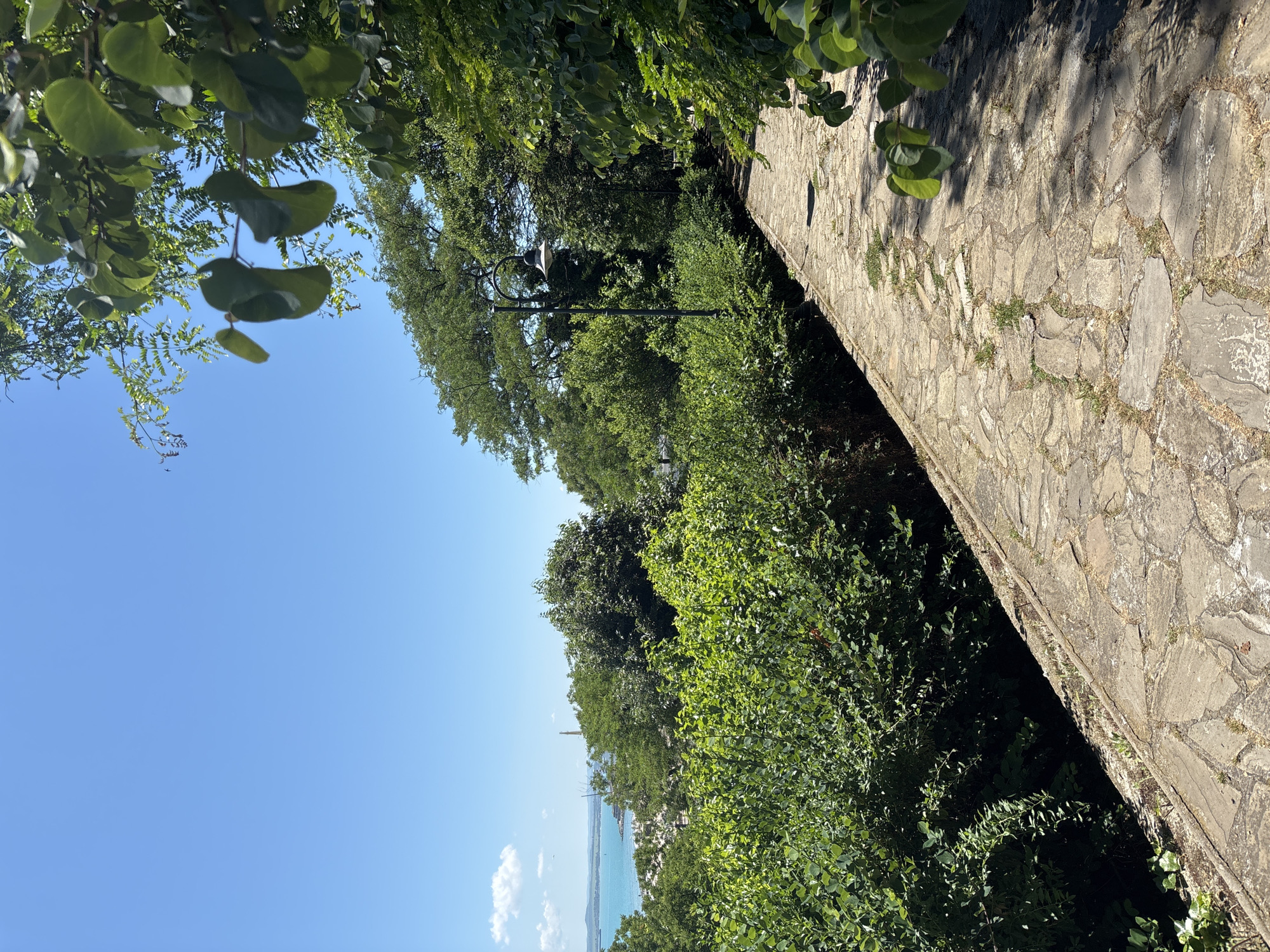 The image depicts a paved stone pathway winding through lush green vegetation. In the background, a clear blue sky stretches over a body of water, likely the Black Sea, with a distant coastline and a few structures visible. The pathway is lined with dense bushes and trees, casting dappled shadows from the sunlight. A street lamp is visible along the path, suggesting it is in a park or recreational area. The scene evokes a sense of tranquility and natural beauty, characteristic of the coastal promenade in Burgas, Bulgaria.