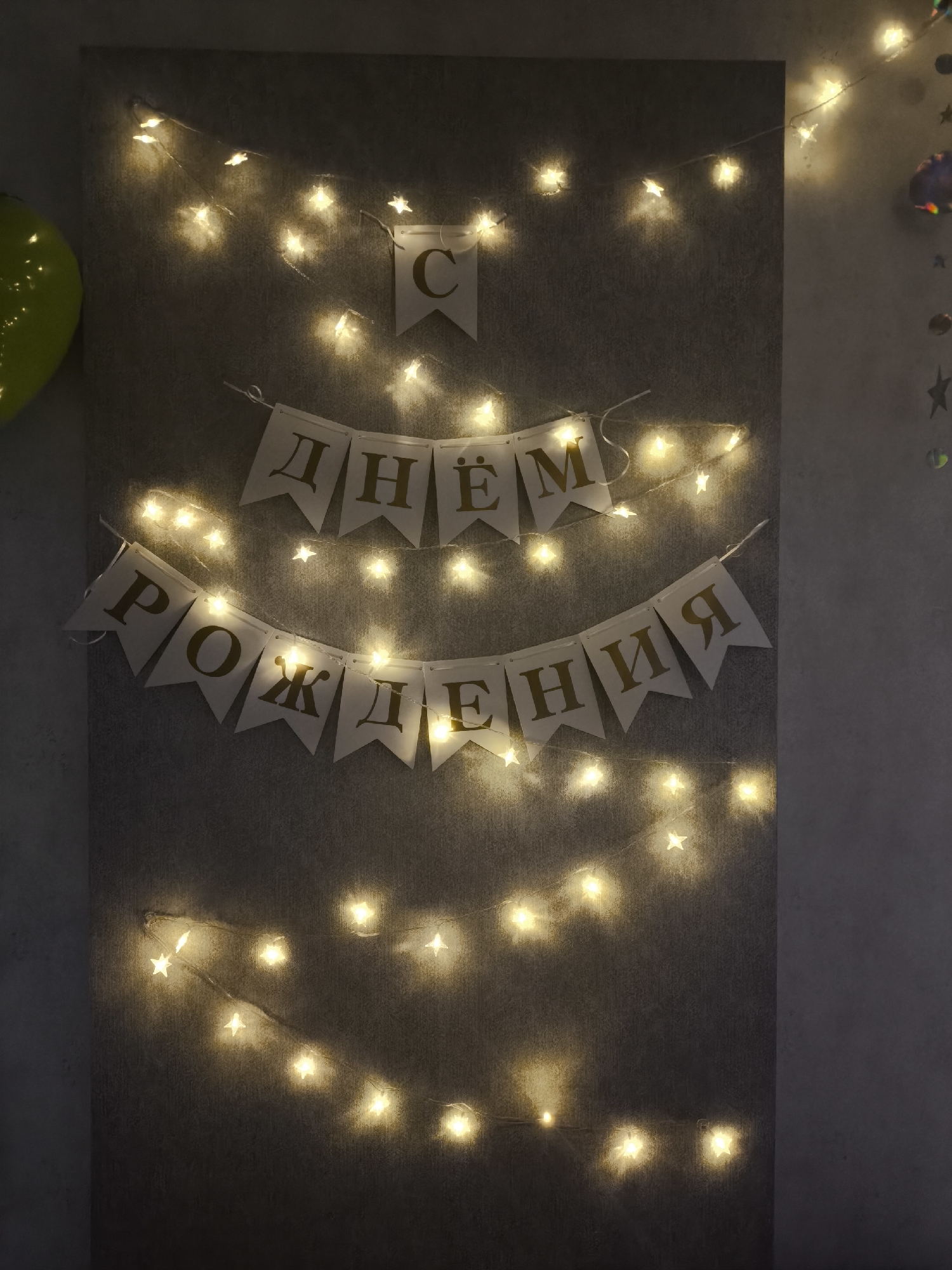 A grey backdrop with star-shaped fairy lights and a banner written in Russian, presumably for a birthday. The banner at the top says "Днём," and the banner in the middle says "Рождения," which translates to "Happy Birthday." There is another banner above the first banner with the letter "С." A green balloon is partially visible on the left. The location is unknown, but the language used in the banner suggests it is most likely located in Moscow, Russia.