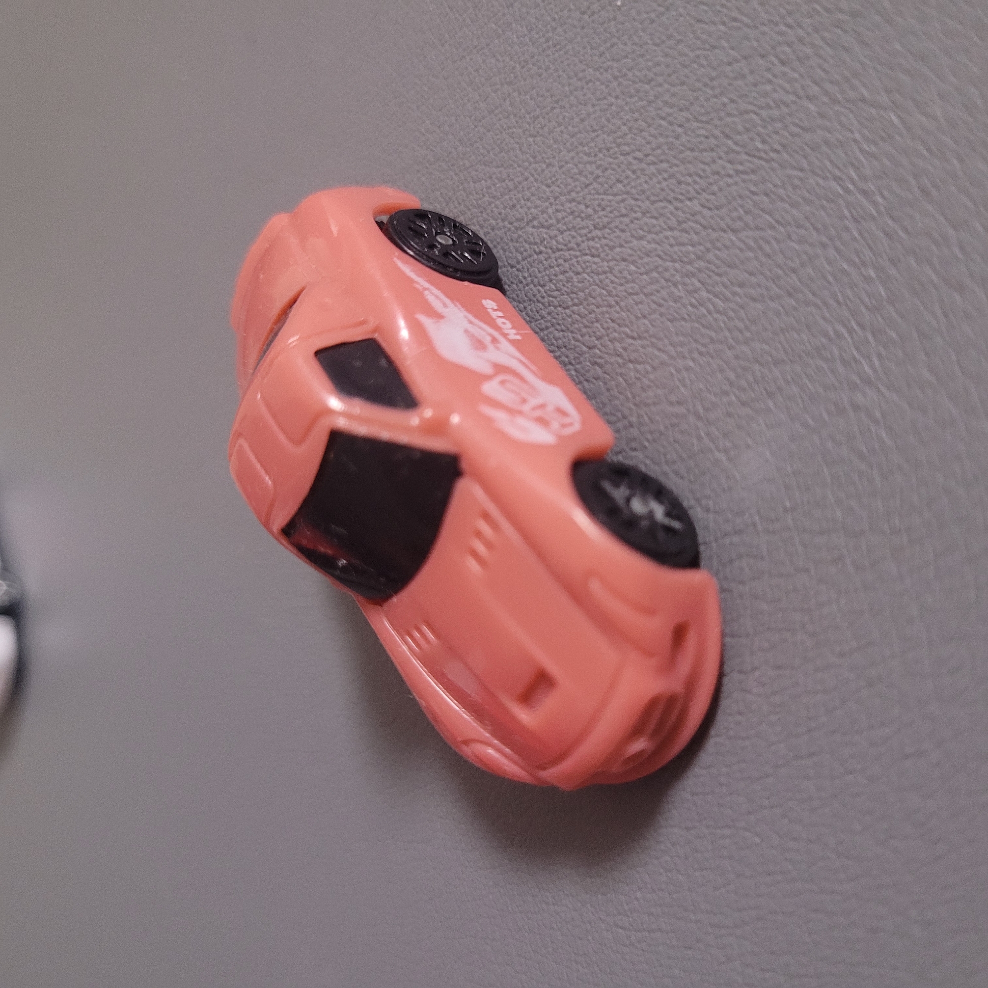 A small, pink toy sports car is on a gray, textured surface. The car has black tinted windows and black wheels. There is a white graphic on the side of the car that appears to be a flame or a stylized animal, with the word "HOTS" in white lettering below it. The car is angled slightly to the right.