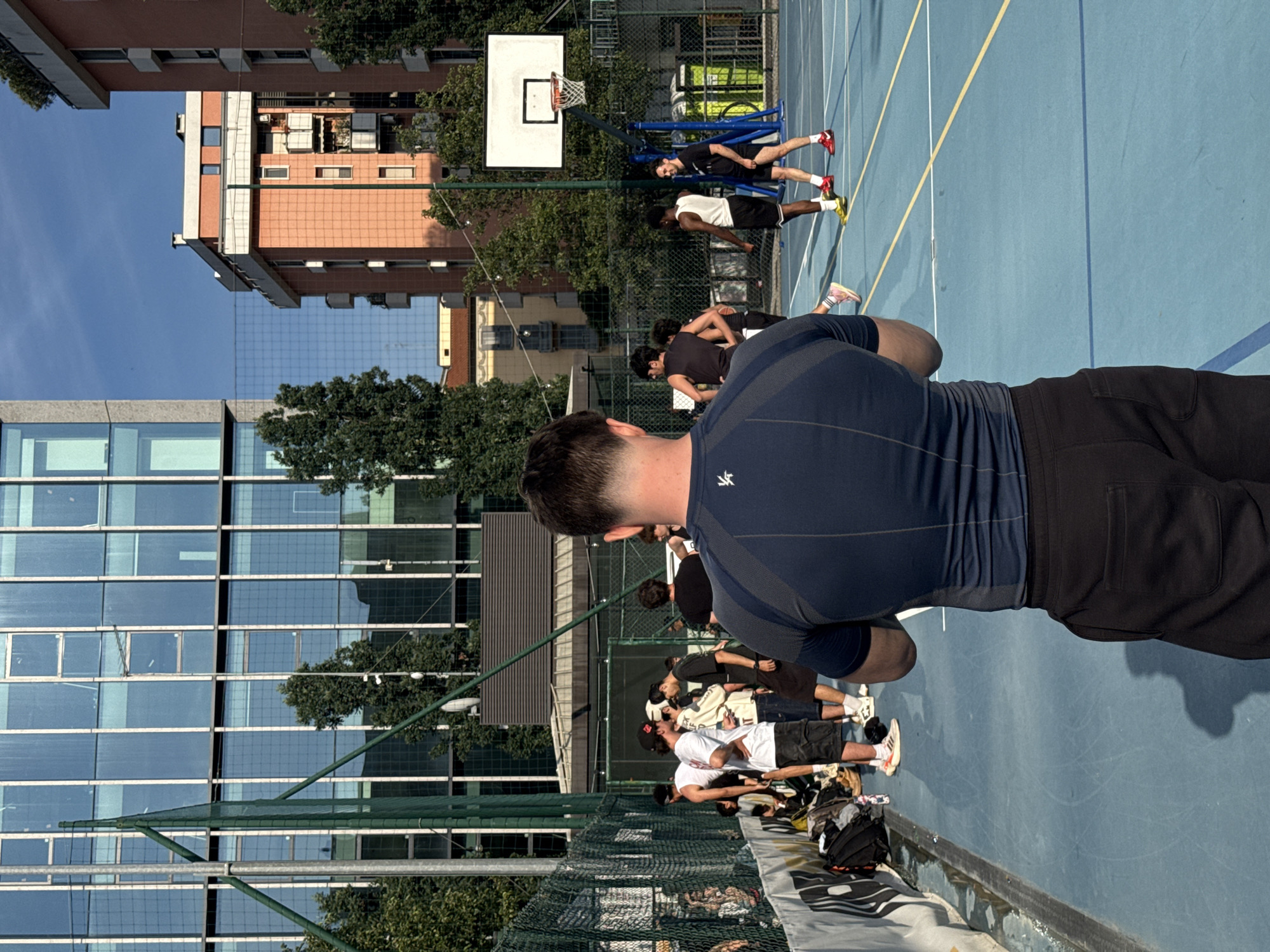 The image depicts an outdoor sports court in an urban area of Milan, Italy, likely within the Porta Nuova, Isola, or Garibaldi district, evidenced by the combination of a modern glass-facade high-rise and traditional terracotta-colored residential buildings in the background. The scene occurs on a clear, sunny day, indicated by a bright blue sky and distinct shadows.
In the immediate foreground, the back of a male subject is visible. He wears a dark blue, short-sleeved athletic top with a white stylized logo on the upper back and black athletic shorts. He stands observing the court.
The court itself is light blue with white and yellow demarcation lines, suggesting a multi-sport configuration. A white basketball backboard, an orange rim, and a white net are prominently featured on the right side of the court. Behind the hoop, two green portable toilets are visible.
Approximately 10-15 men are distributed across the scene. On the left side of the court, a group of 6-8 men in various athletic and casual wear (t-shirts, shorts, sneakers) congregates near a green chain-link fence that borders the court. Personal belongings, including bags and clothing, are arranged on the ground beside the fence. On the right side of the court, near the basketball hoop, 4-5 additional men in athletic attire are standing. One individual in a black t-shirt and red sneakers smiles towards the left side of the frame. The subjects appear to be either playing basketball, resting, or preparing for a game.
The background features a diverse urban landscape: a tall, multi-story modern building with a reflective blue glass and white-framed facade dominates the left skyline. To the right, multiple residential buildings constructed of terracotta-colored brick or stucco, with white trim and balconies, are visible. Green trees are interspersed throughout the background, separating the court from the distant buildings.