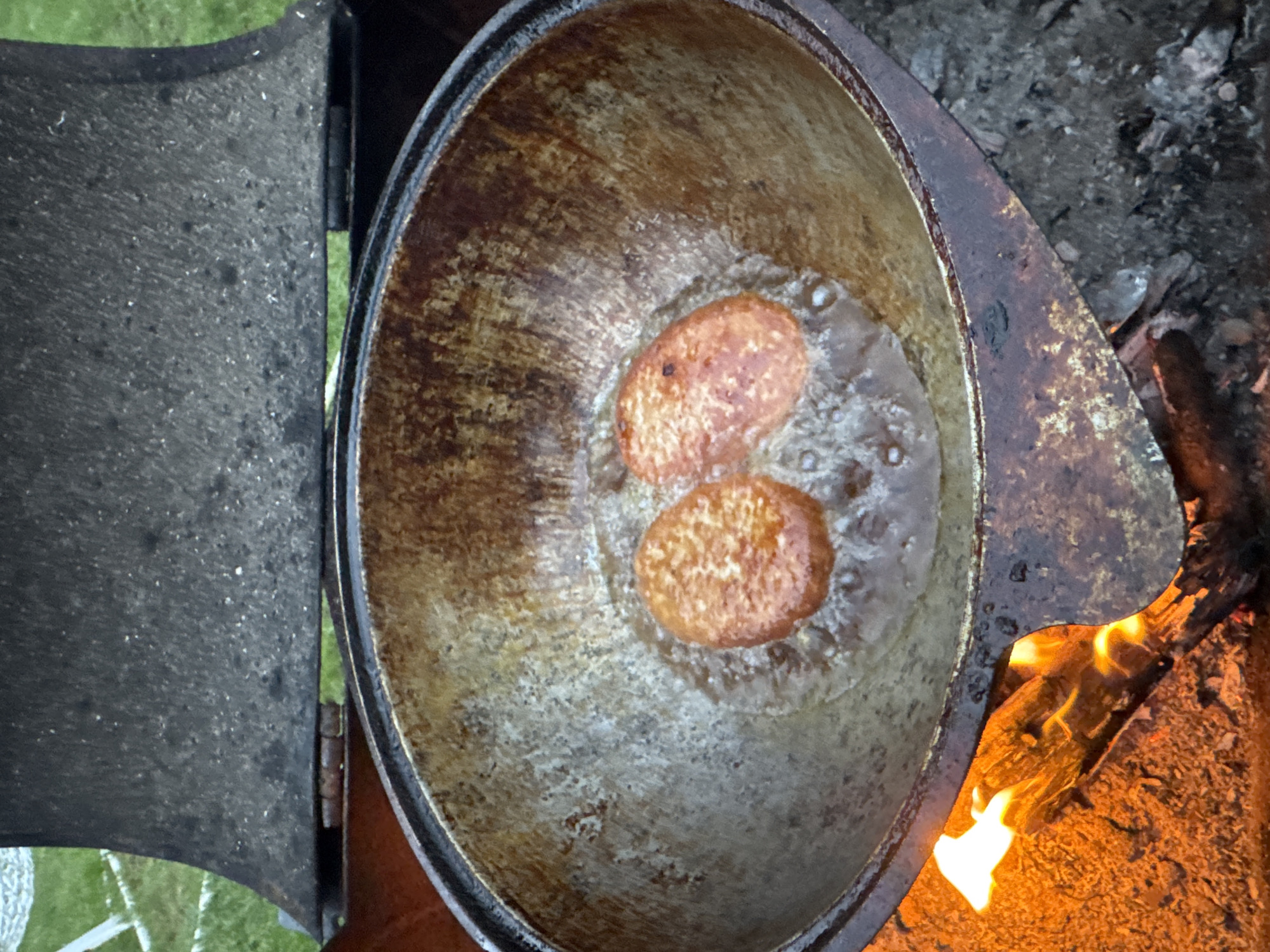 A top-down shot shows two fried patties cooking in oil in a weathered metal pot positioned over an open fire. The pot sits atop a stove with a partially open lid to the left. To the right, flames rise from the fire, with ash and charred wood nearby. A patch of grass is visible in the lower left corner. The image was taken in the city of Chaikovsky, Russia.