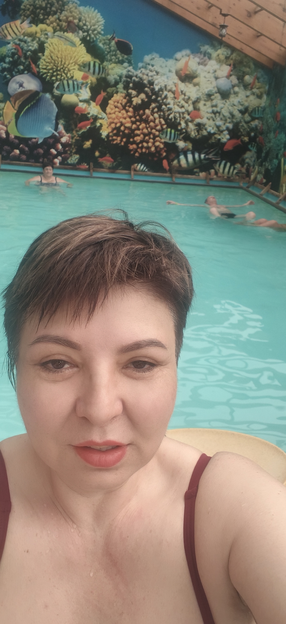 A woman with short, damp, dark brown hair, wearing a dark red swimsuit top, takes a close-up selfie while in a swimming pool. She looks directly at the camera with a neutral expression, her lips slightly parted, and light coral lipstick visible. Water droplets are present on her skin, particularly on her chest and neck.
The background reveals a vibrant indoor swimming pool environment. The pool water is clear and light blue. The walls surrounding the pool are adorned with a large, detailed mural depicting an underwater coral reef scene. The mural features various types of colorful corals in oranges, yellows, and browns, interspersed with numerous fish, including striped varieties, red fish, and a larger butterflyfish-like creature.
Several other people are visible in the pool, relaxing or swimming. One person with light hair is seen floating on their back, and another individual is standing in the water further back. The overhead structure indicates an indoor setting, with wooden beams visible. The overall scene suggests a leisure activity in a themed swimming facility, located in Votkinsk, Russia. There is no visible text in the image.