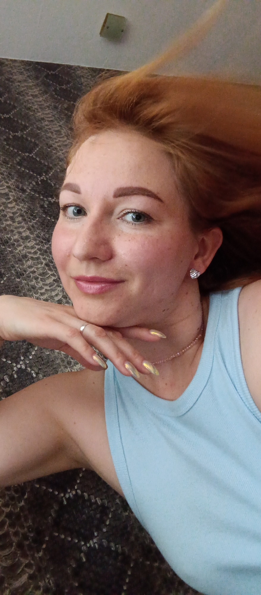 A fair-skinned woman with red hair is lying down, resting her chin on her hand. She is wearing a light blue tank top and a delicate pink necklace. A silver ring is visible on her finger, and she has heart-shaped earrings. Her fingernails are long and have a metallic finish. She is looking directly at the camera with a slight smile. The background includes a textured, dark-colored surface, potentially a blanket or rug, and a light-colored wall with a light fixture. The location is unknown.