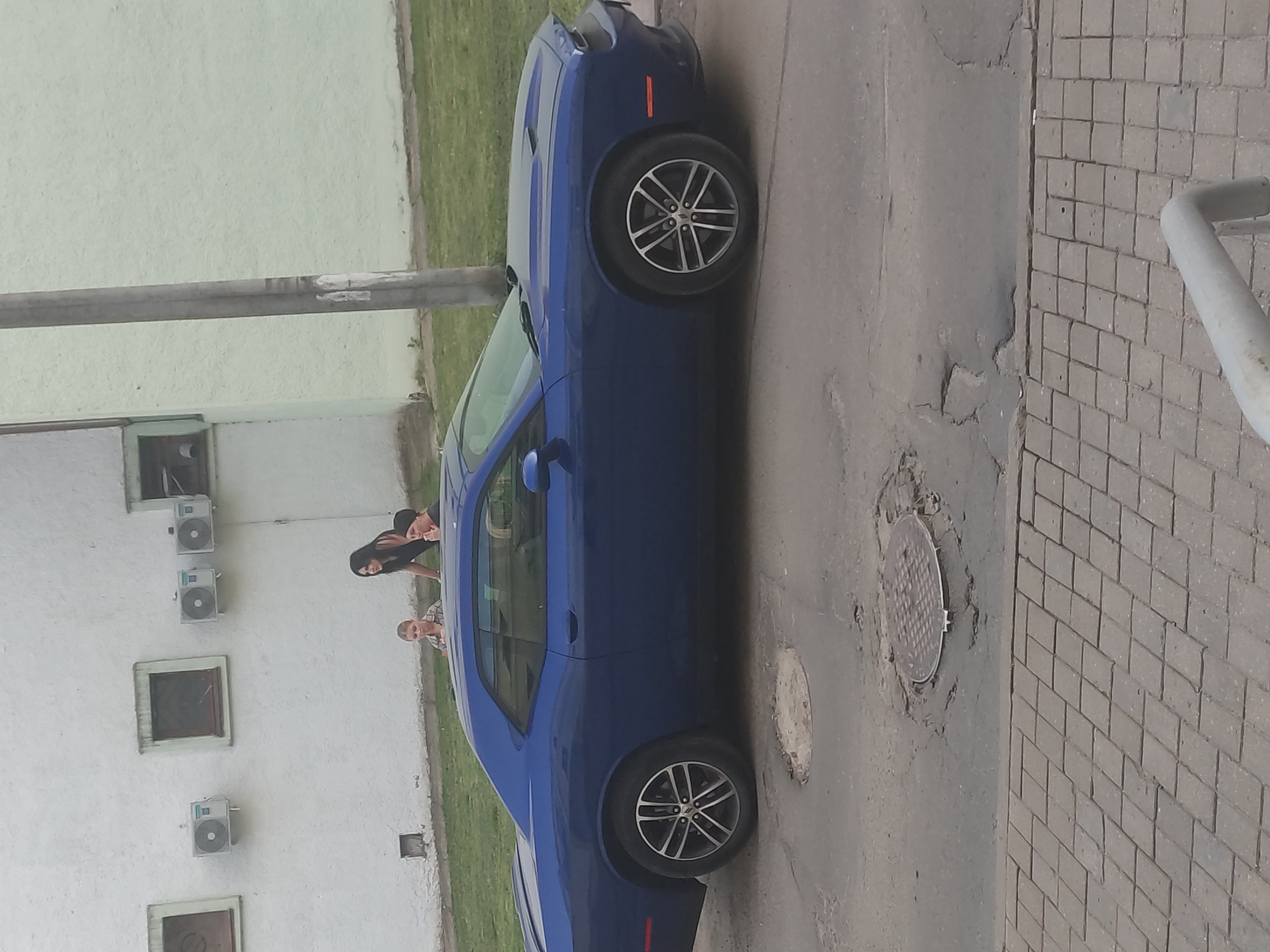 In Mahilyow, Belarus, a blue Dodge Challenger is parked on a paved surface with visible potholes and a manhole cover. Two young children, a girl with blonde hair and a boy, are standing near the car. A woman with dark hair is leaning over the car, interacting with the children. In the background, a white building with multiple windows and air conditioning units is visible, adjacent to a light green building. A utility pole stands near the vehicles. The scene appears to be an urban street or parking area.