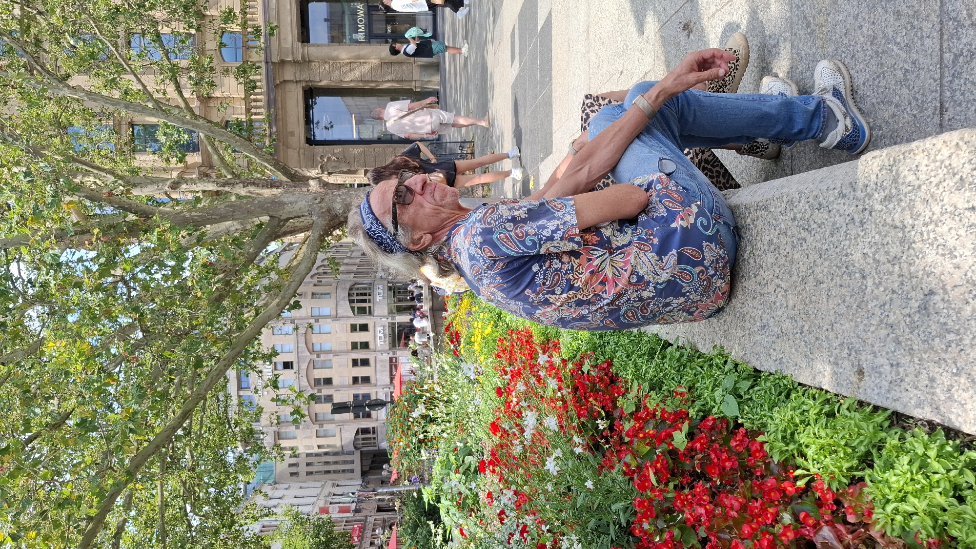 The image shows an elderly Caucasian man with grey hair, wearing a blue bandana, sunglasses, and a patterned blue shirt, sitting on a stone bench in a city square. He is looking to his right. In front of him, there is a flower bed filled with red and white flowers. Behind him, there are buildings, a large tree, and other people walking in the background. The scene appears to be a sunny day. The architecture and the presence of a "RIMOWA" store indicate the location is in Cologne, Germany, likely near the Schildergasse shopping street.