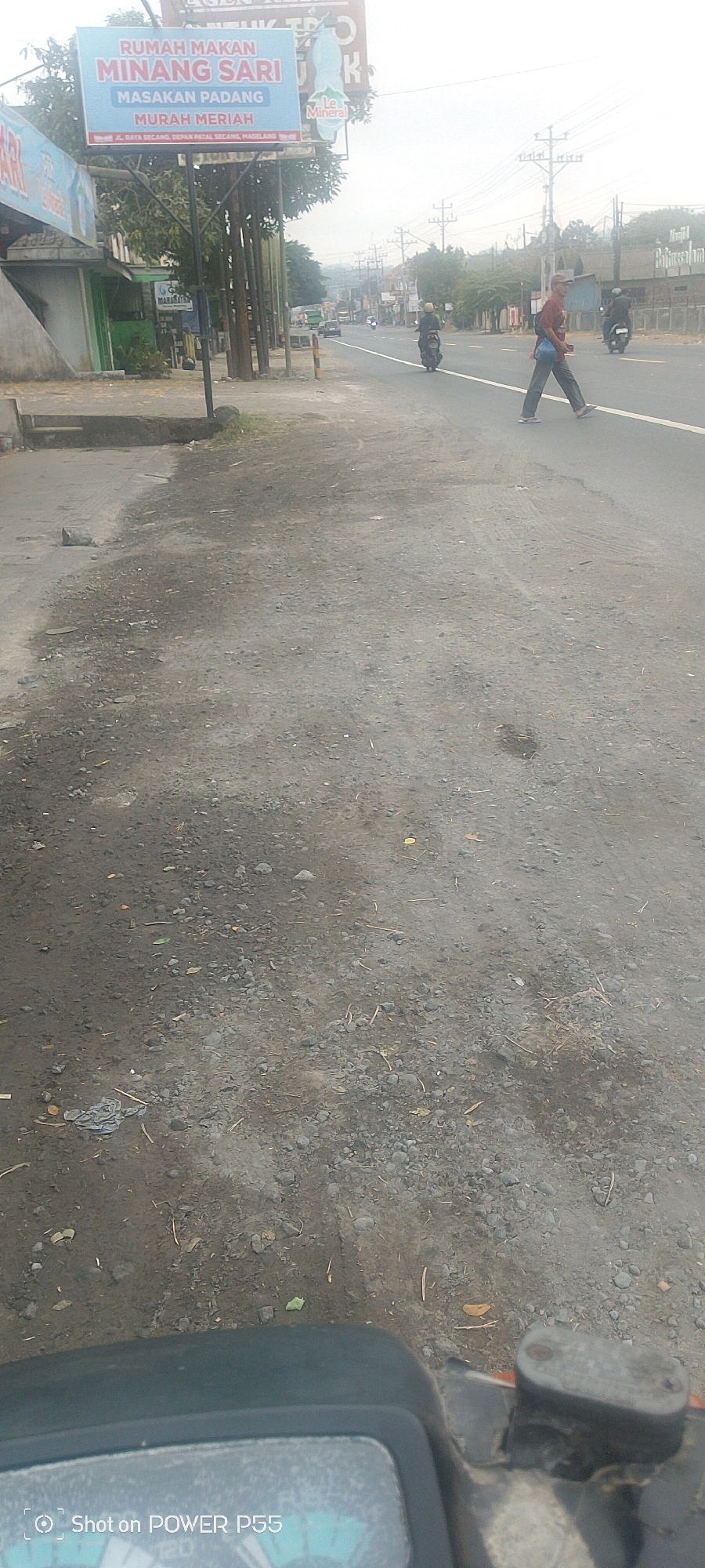 The image depicts a street scene, likely in a city. A prominent billboard for "RUMAH MAKAN MINANG SARI" (Minang Sari Restaurant) is visible on the left, advertising "MASAKAN PADANG MURAH MERIAH" (Affordable Padang Cuisine). The street itself is paved, with a dirt shoulder to the left. Several utility poles line the side of the road. Traffic is present, with motorcycles and cars visible. A person is crossing the street in the middle ground, wearing dark pants and a light-colored shirt, carrying a bag. Another person on a motorcycle is visible further down the road. The environment appears to be urban or semi-urban, with a slightly hazy sky. The foreground shows the dashboard of a vehicle, likely a motorcycle, with a digital display.