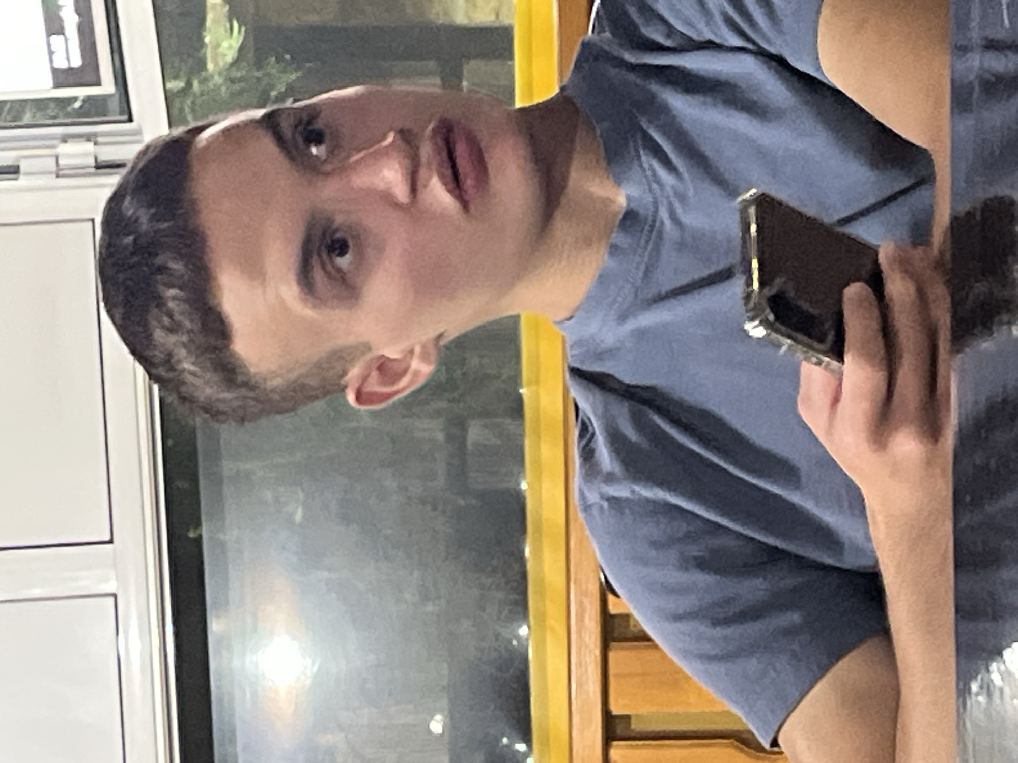A forensic examination of the image reveals a close-up, waist-up view of a young adult male seated indoors in what appears to be a commercial establishment within the city of Basarbovo, Bulgaria. The male, with short dark hair and fair skin, is observed looking towards the viewer's left with his mouth slightly agape, suggesting an expression of surprise or contemplation. He is wearing a plain, crew-neck, short-sleeved blue t-shirt. His right hand holds a dark-colored smartphone, possibly with a clear protective case, while his left arm rests on a dark, reflective table surface in the foreground.
The background features a large window, behind which blurred lights and faint dark shapes, indicative of outdoor foliage or structures at night, are visible. Above the window are white paneled structures, possibly parts of an interior wall or ceiling, featuring dark vertical lines suggesting seams or frames. Below the window, a prominent yellow horizontal bar or ledge is present, beneath which a wooden structure, possibly a bench or booth backing, extends to the left. The overall lighting suggests an interior setting, likely evening or night due to the illuminated reflections in the window.