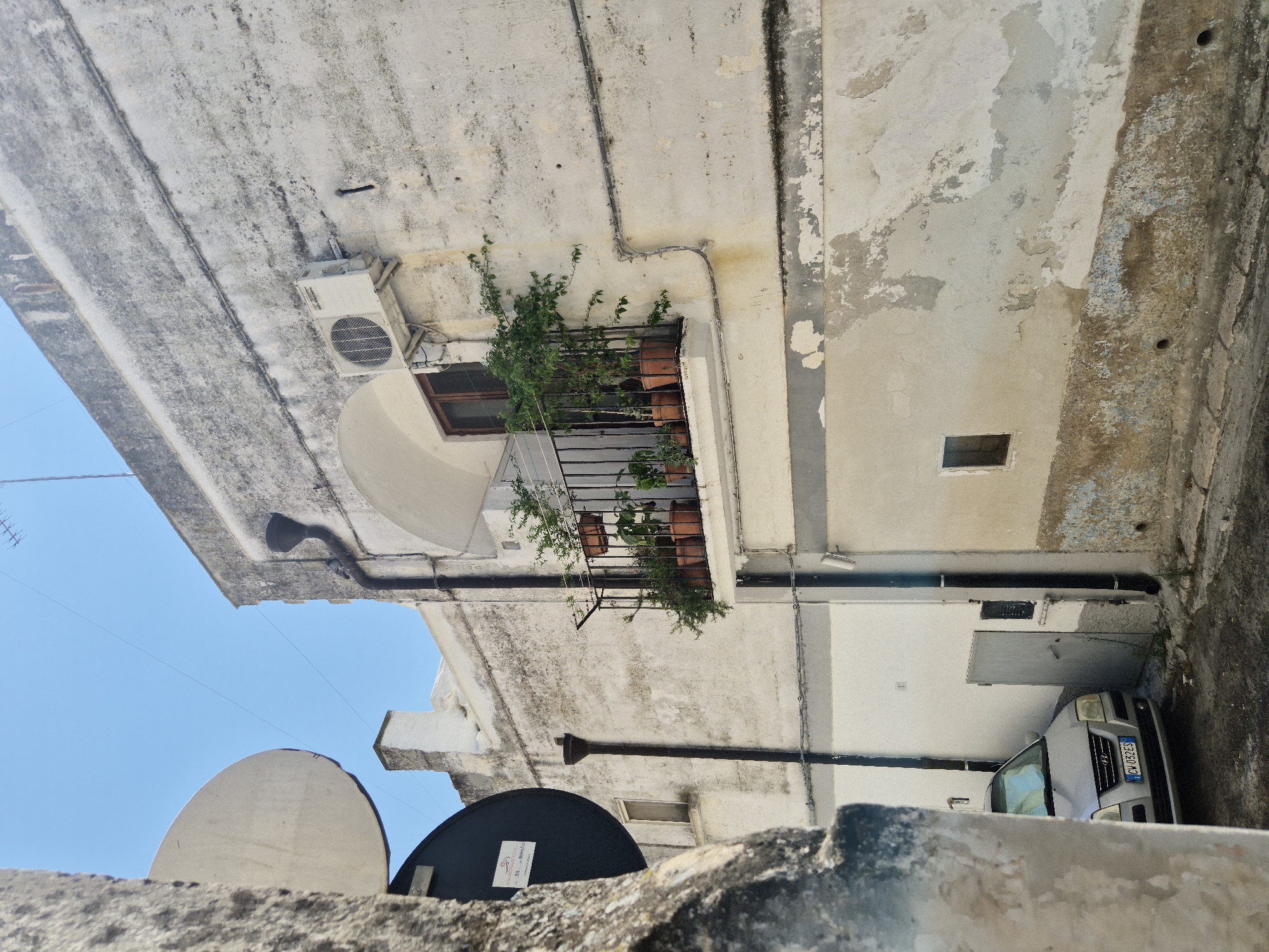 In a narrow alleyway in Laterza, Italy, a weathered white building with peeling paint dominates the scene. A small balcony, adorned with potted plants, juts out from the upper floor. An air conditioning unit is mounted on the wall above the balcony. To the left, a portion of a large, circular traffic sign is visible, partially obscured by a rough-textured stone pillar. A silver car is parked in the alleyway, with its license plate reading "CW 052ES". The ground is paved with asphalt, and the sky is a clear blue.