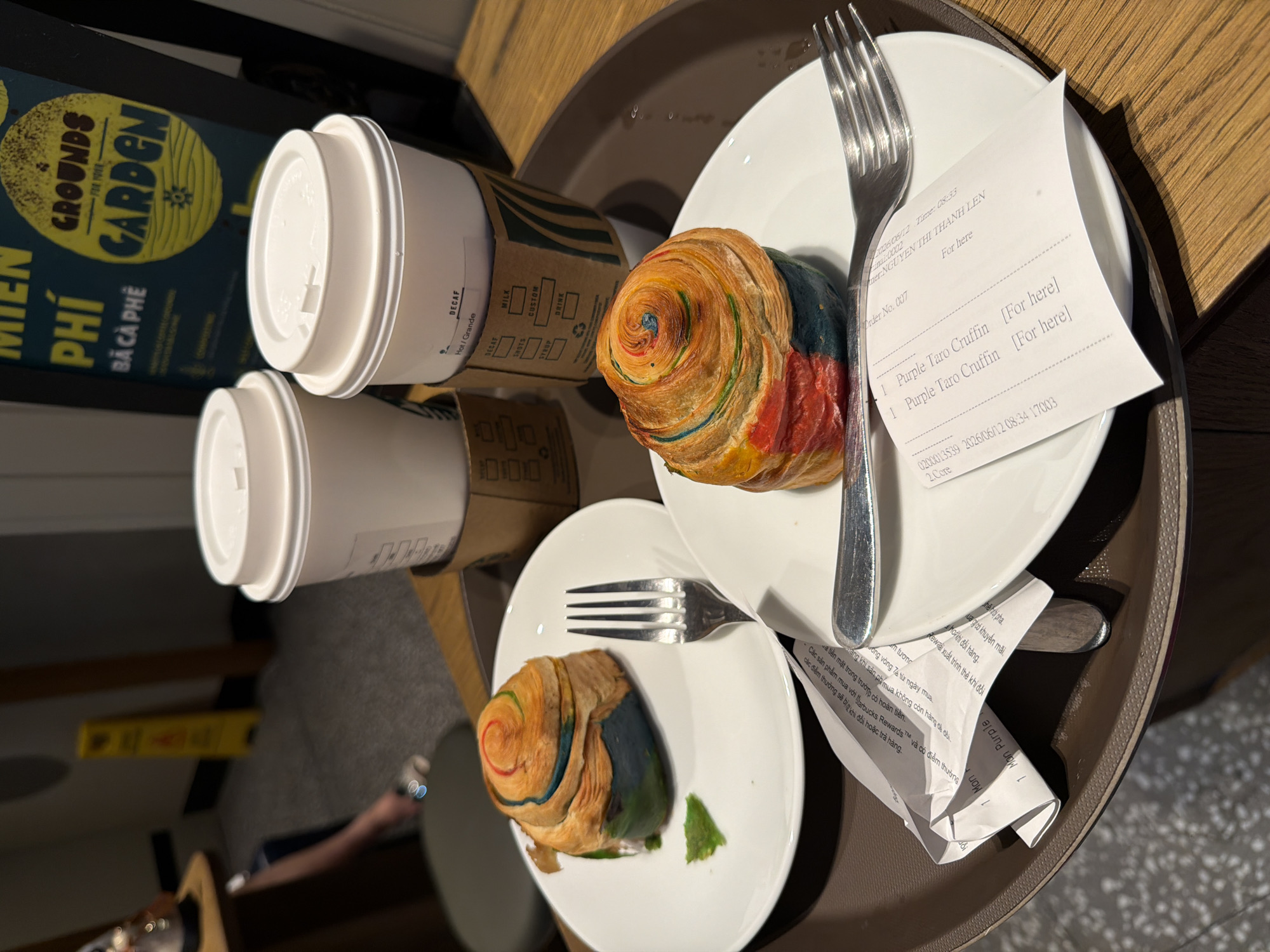 The image shows a tray on a wooden table in what appears to be a cafe in Da Nang, Vietnam. On the tray are two white paper cups with cardboard sleeves and two colorful pastries on white plates, each with a fork. One pastry is a rainbow-colored cruffin with visible layers of dough, and the other is similar in appearance but slightly obscured. There are also receipts on the plates. In the background, a sign with Vietnamese text and "GROUNDS FOR YOUR GARDEN" is visible. A yellow safety cone is also in the background. The overall scene suggests a casual meal or break at a cafe.