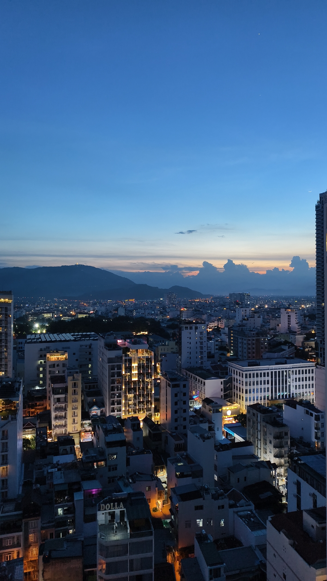 This is a drone shot of Nha Trang Ward, Vietnam, taken at dusk. The camera is positioned high up, offering a panoramic view of the city. In the foreground, several buildings of varying heights and architectural styles dominate the frame. Some are modern, with clean lines and illuminated windows, while others appear older, with intricate details. The light from the windows suggests that the city is coming alive as night falls.
In the middle ground, a dense urban landscape stretches out, with more buildings and streetlights creating a warm glow. In the background, a range of mountains forms a silhouette against the colorful twilight sky. The sky itself is a gradient of deep blue at the top, transitioning to hues of orange and pink near the horizon, where the sun has recently set. A few wispy clouds are scattered across the sky, catching the last rays of sunlight. The overall impression is one of a vibrant city bathed in the soft light of dusk, with a natural landscape providing a dramatic backdrop.
