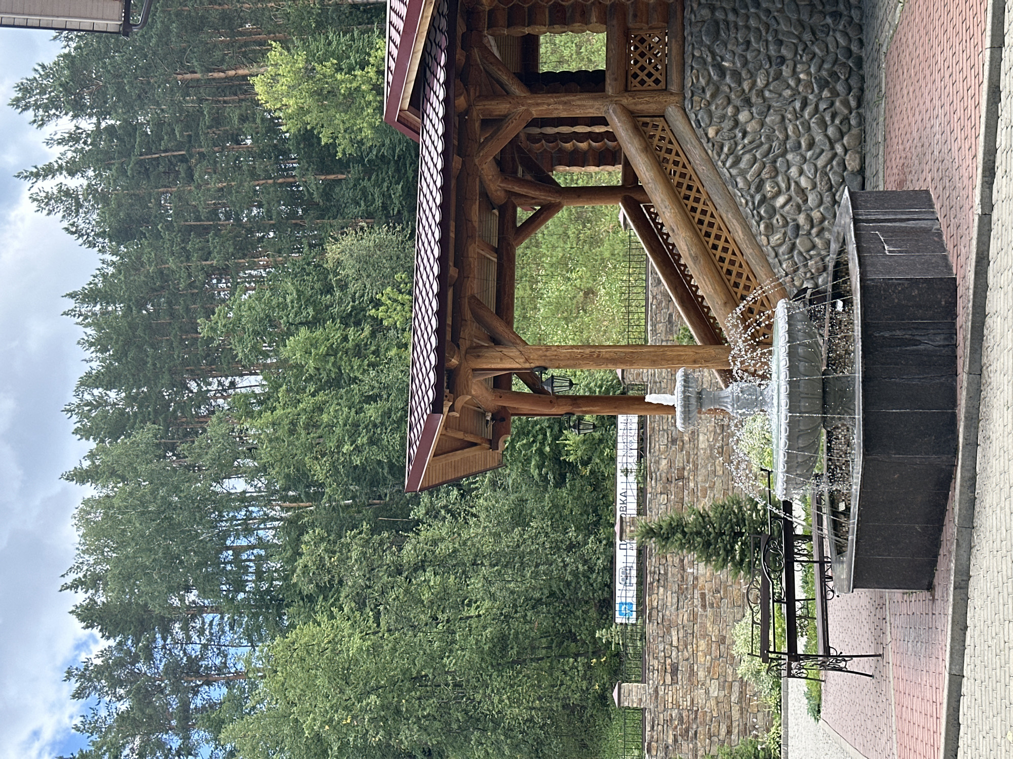 A stone wall with a black metal fence sits in the foreground. Behind the wall is a large wooden building with a red tile roof and a stone base. A wooden staircase leads up to the building's entrance. A multi-tiered fountain with water spraying into the air is in front of the building, on a paved area. A dark metal bench with decorative scrollwork is to the left of the fountain. A small evergreen tree stands behind the bench. In the background, a dense forest of pine and deciduous trees covers a hillside. The sky is cloudy. The scene is in Belokurikha, Russia.