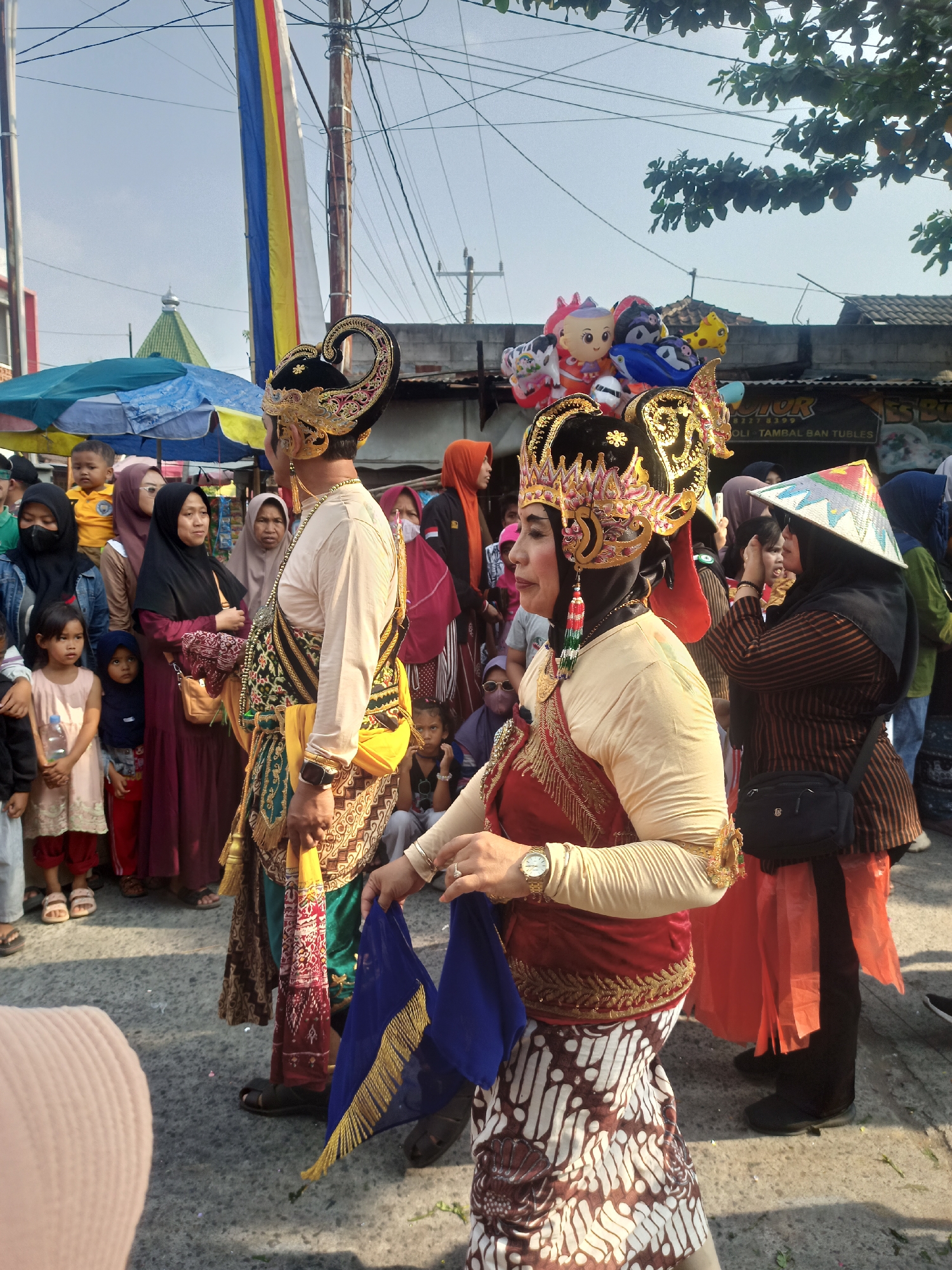 The image depicts a street scene in Saren, Indonesia, likely during a festival or parade. In the foreground, two individuals are dressed in traditional Javanese attire. The person on the left wears a cream-colored long-sleeved shirt, a gold and black headdress adorned with red and gold embellishments, and a patterned cloth wrapped around their waist and legs. They hold a blue fabric with gold fringe. The person on the right wears a similar cream-colored top, a large, ornate gold headdress with red and gold detailing, and a red vest with gold embroidery. Their lower garment is a batik patterned skirt.
Behind them, a crowd of onlookers watches. Many are wearing headscarves, and some children are visible. Some individuals are carrying balloons in the shape of cartoon characters. The environment appears to be an outdoor market or street with temporary stalls and buildings visible in the background, including tarpaulin canopies. Power lines and a tall pole with colorful vertical banners are also present. The ground is paved with asphalt. The lighting suggests it is daytime with a clear sky.