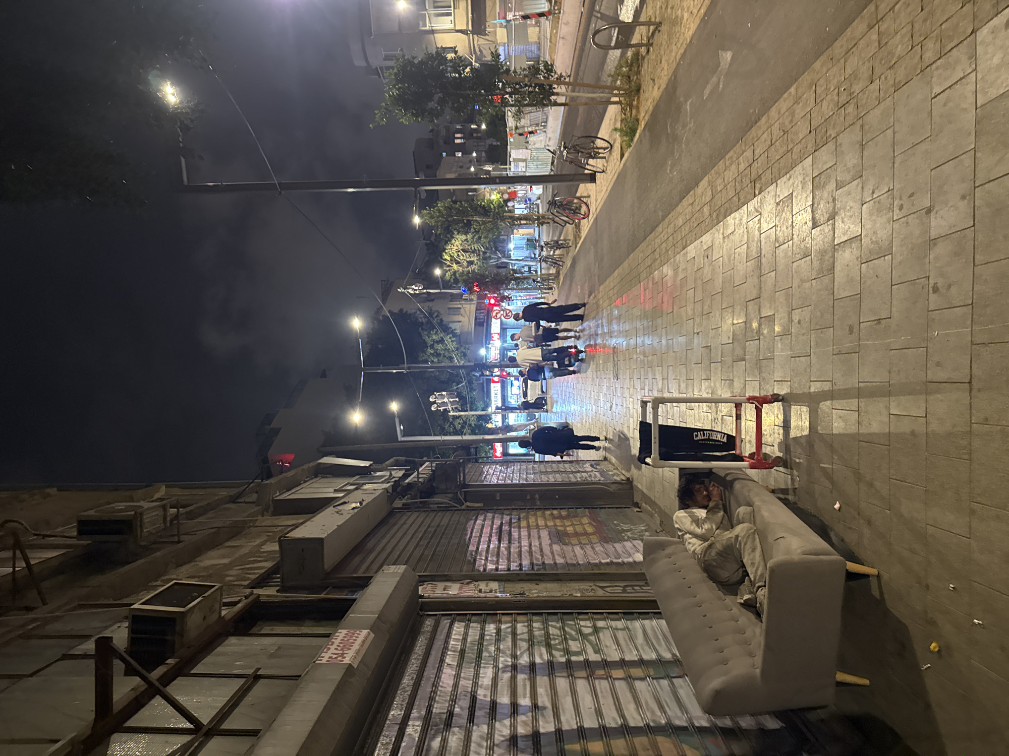A wide-angle, nighttime shot captures a street scene in Tel Aviv, Israel. Along the right side of the frame is a sidewalk with cobblestone pavers. A gray couch sits on the sidewalk, occupied by a man lying down. His legs stretch toward the sidewalk's edge, and he appears to be using his phone. Behind the couch, against the wall of a building, is a metal and red structure with a black bag labeled "CALIFORNIA."
The left side of the frame features commercial buildings with closed, corrugated metal security gates. Air conditioning units are visible on the rooftops of these buildings. The street extends into the distance, illuminated by streetlights, with pedestrian traffic visible. Several bicycles are parked along the sidewalk further down the street. The sky above is dark, with clouds obscuring the upper reaches of the buildings and streetlights.