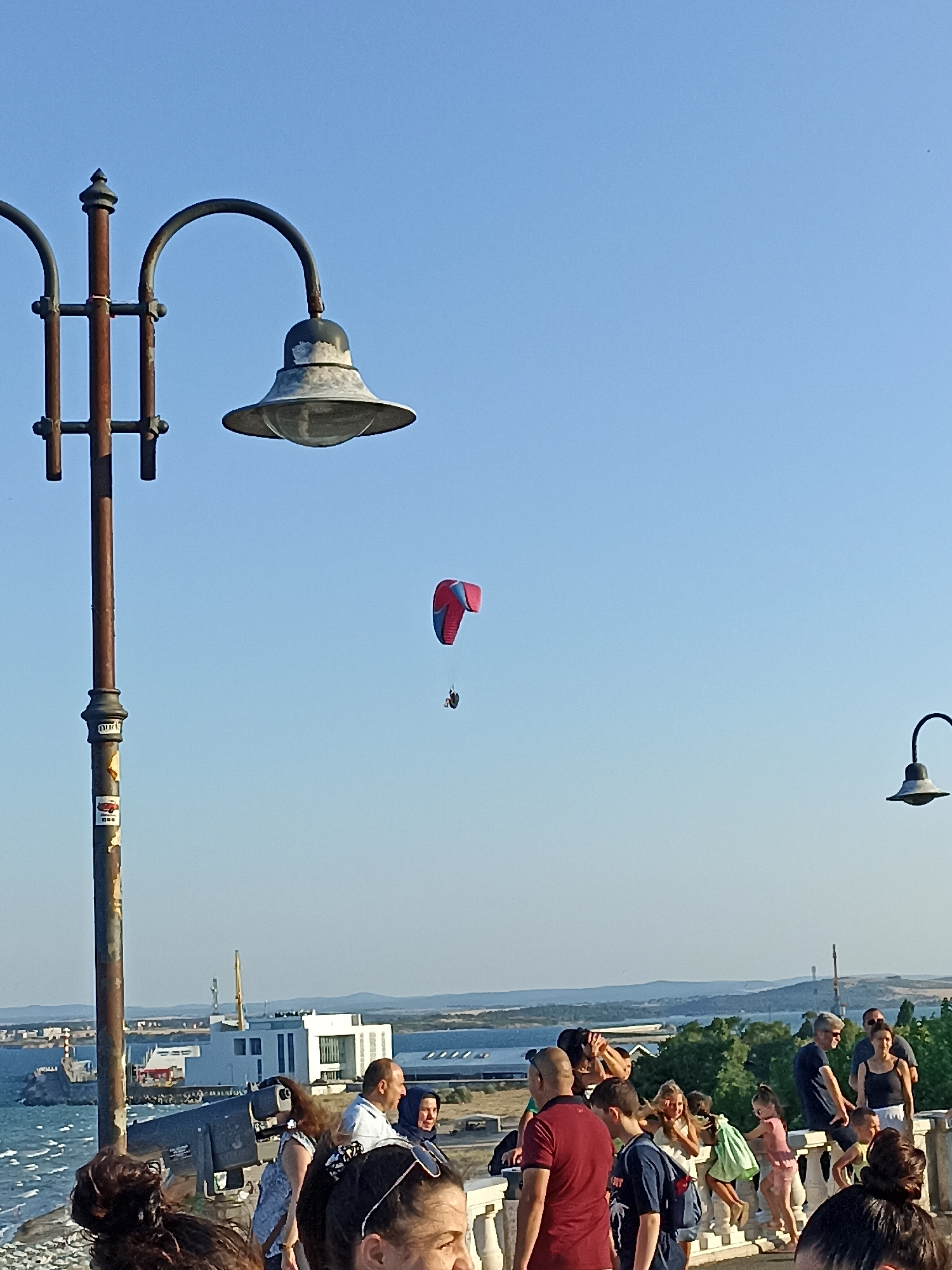 The image captures a panoramic view of the Burgas Sea Garden with a clear blue sky. In the foreground, a lamppost with a rustic, weathered finish stands tall, adorned with stickers. A group of people are visible, some appearing to be tourists and locals enjoying the scenery. Among them, a paraglider is seen gracefully suspended in the air, adding a dynamic element to the scene. In the background, the expansive sea stretches towards the horizon, with ships and buildings lining the coast, hinting at the bustling port city of Burgas. The overall atmosphere is one of a vibrant, outdoor recreational space, bathed in the warm glow of sunlight.