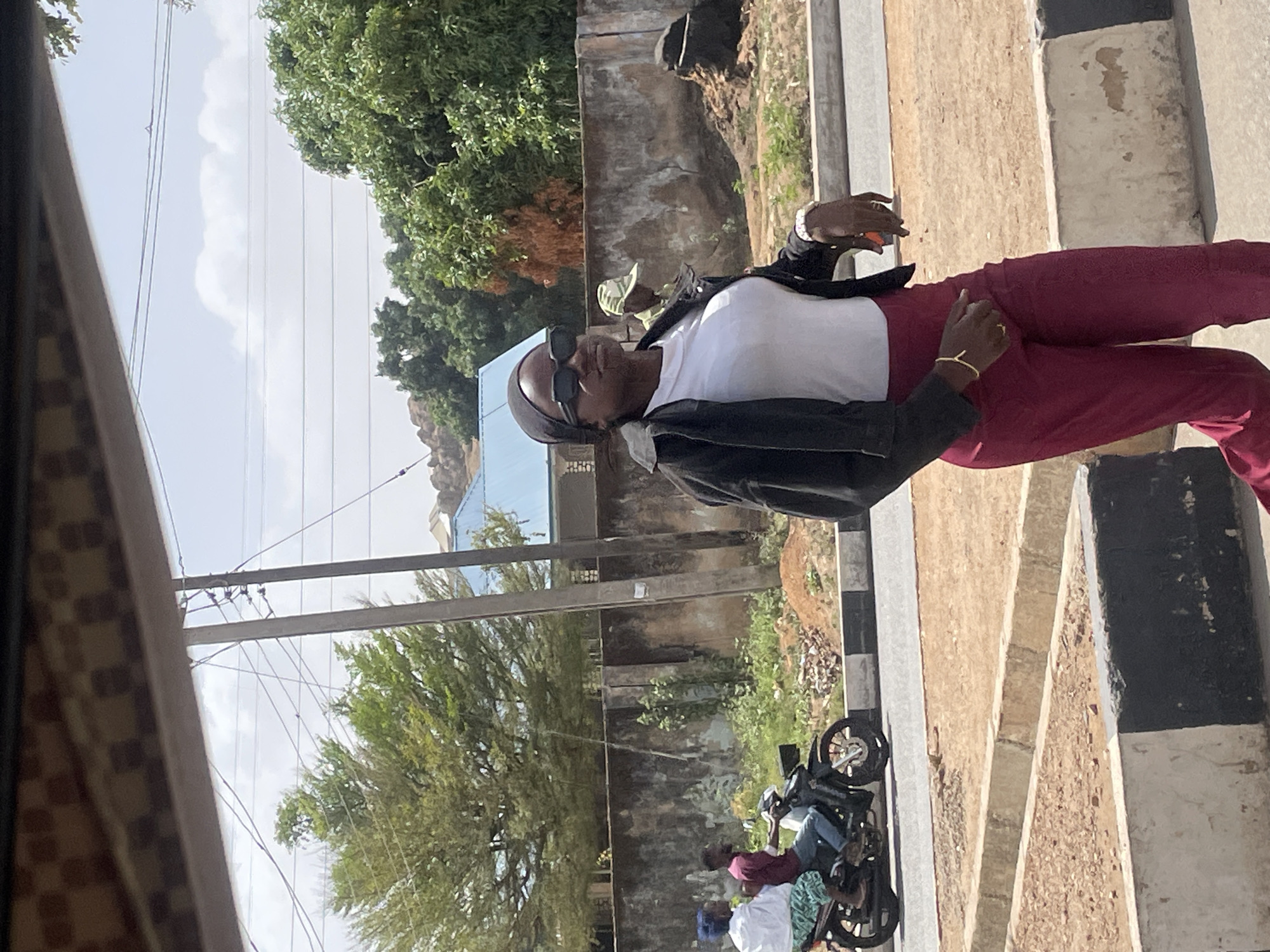 A woman wearing a headscarf, sunglasses, a white shirt, and a black jacket, along with maroon pants, stands on a sidewalk. The location is Gura Top, Nigeria. Behind her is a stone wall and a street scene with parked motorbikes and other people. Power lines are visible overhead. A partial view from inside a vehicle frames the left side of the image.