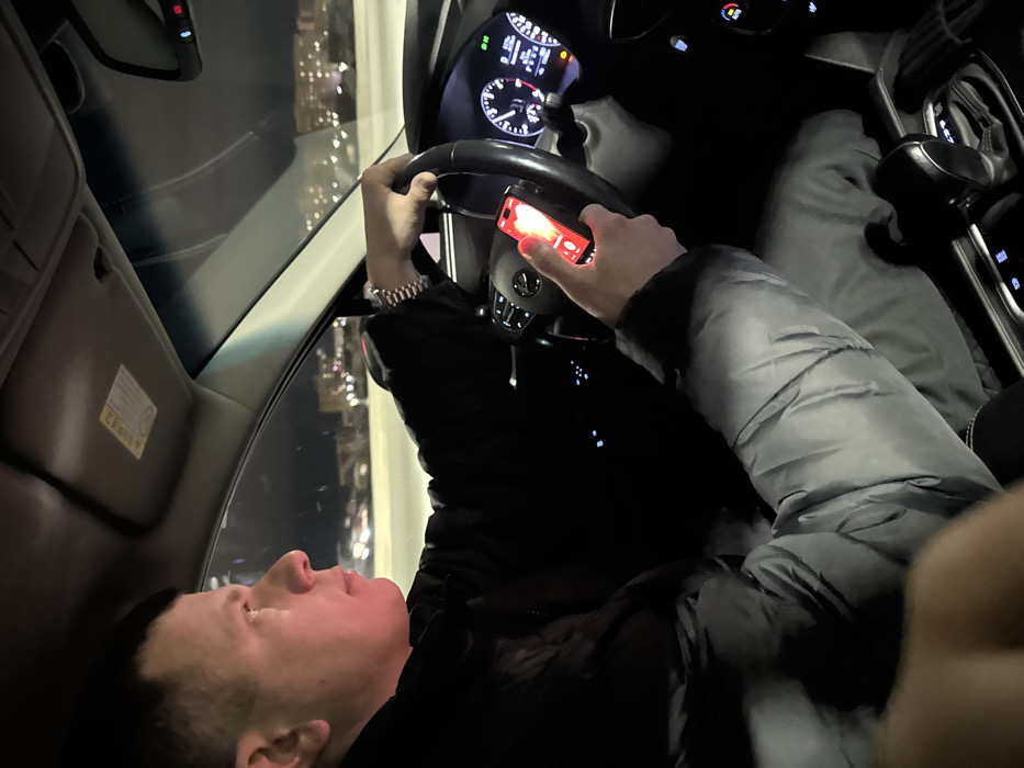 A person is inside a car, looking up and towards the left. They are wearing a black jacket and a light grey puffer jacket over it, as well as a watch. The person is holding a phone with a red case in their right hand on the steering wheel. The car's dashboard and a night cityscape are visible through the windshield.