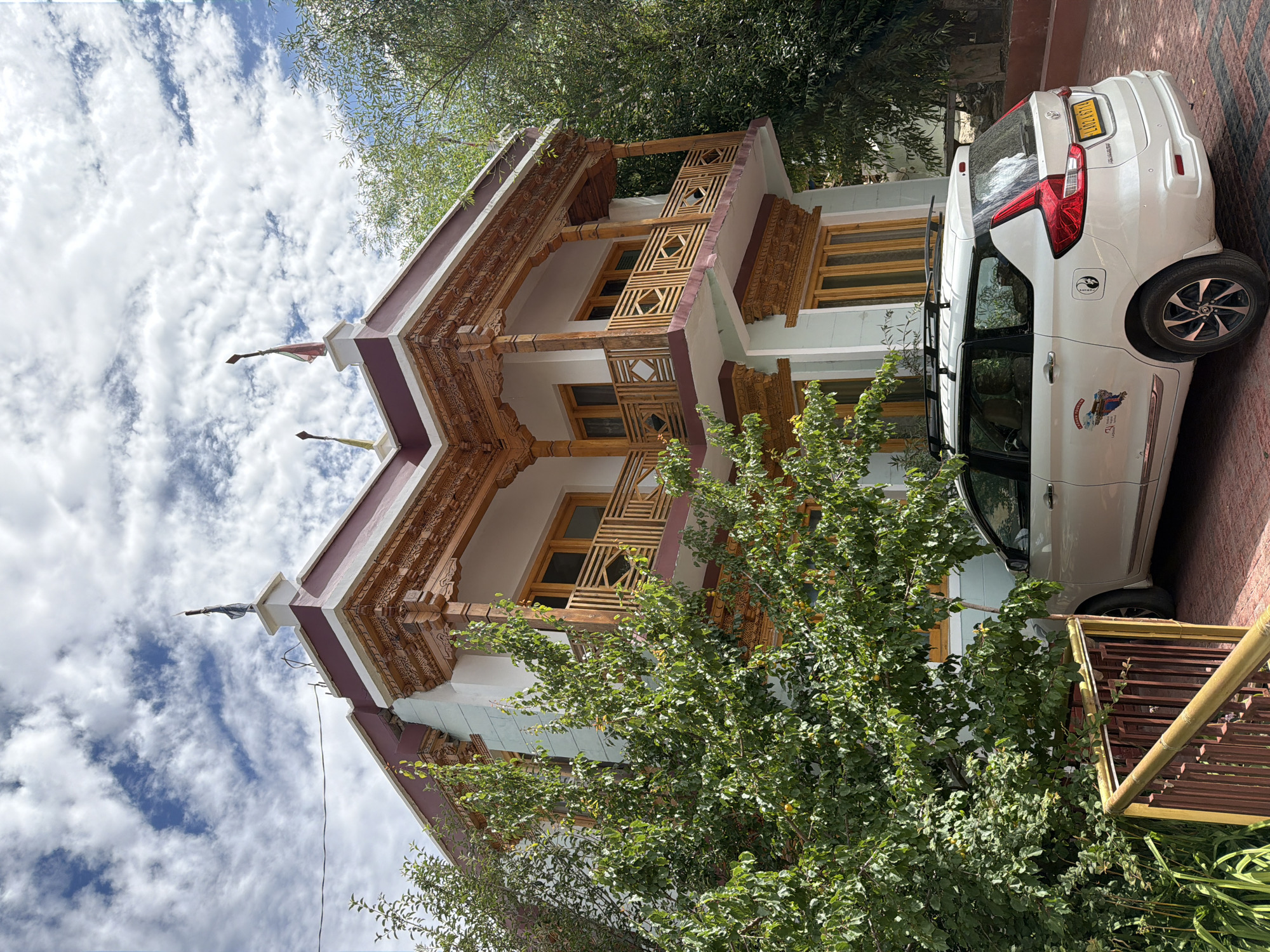 In Leh, India, a white multi-purpose vehicle is parked in front of a light blue and white building with ornate wooden architectural details and a maroon trim. The vehicle has a roof rack, a license plate reading "LA07A 9714", and several stickers on its rear passenger door, including one depicting mountains and another with a yin and yang symbol. To the left of the vehicle, a lush green tree with yellow fruits partially obscures the building. A yellow and brown railing separates the parking area from a small patch of greenery. The sky above is filled with dramatic white clouds against a bright blue backdrop.