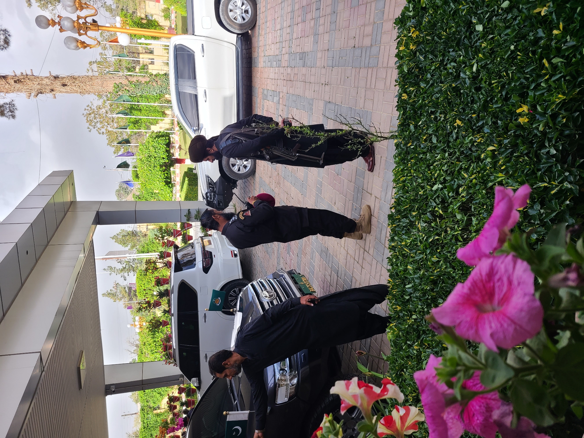 The image shows a scene outside a building in Quetta City Tehsil, Pakistan. Three men are present. Two of the men are wearing dark uniforms and berets and carrying rifles. The third man is wearing black traditional clothing. They are standing near a row of parked vehicles, some of which have Pakistani flags attached. A flower bed and decorative shrubbery are also visible. In the background, there's a tree and a lamppost.