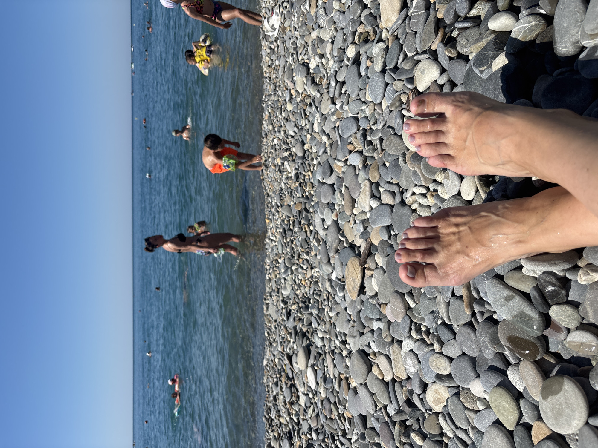 The image depicts a sunny day at a pebbly beach, likely in Sochi, Russia, given the visual cues of a Black Sea coastline. The foreground is dominated by a dense field of smooth, grey and multi-colored pebbles. In the lower center of the frame, a person's bare feet are visible, with wet skin indicating recent immersion in water. The feet are positioned facing away from the camera, towards the sea.
The middle ground shows the shallow edge of the sea, where several people are present. A woman in a black bikini stands in the water, facing towards the right of the frame. Further out in the sea, other individuals are visible, some swimming and others on inflatable toys. Notably, a young boy in orange shorts is bent over near the water's edge, holding what appears to be a green object. A child in a yellow flotation device is also in the water.
The background features a vast expanse of calm, blue water under a clear, bright blue sky. Several boats or jet skis are visible in the distance, suggesting recreational activities. The overall impression is one of a typical summer day at a popular beach resort.