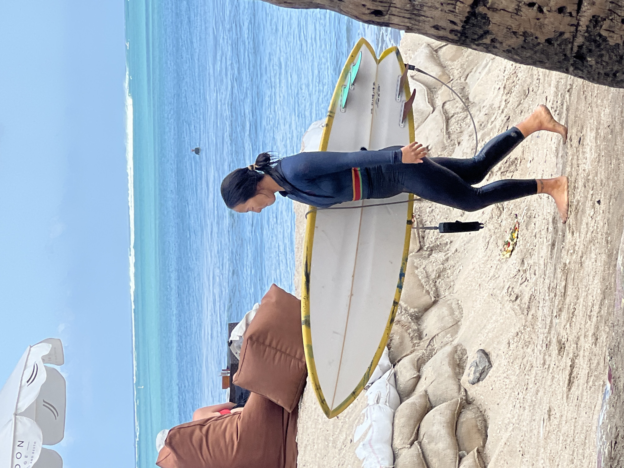 A woman in a dark blue wetsuit is walking on a sandy beach in Kuta, Indonesia, carrying a white surfboard with yellow accents. She is wearing her hair in a ponytail and has a red and green stripe around her waist. The surfboard has three fins. In the background, the ocean is a bright turquoise color with waves breaking in the distance. There is a line of sandbags on the beach behind the woman, and further back, a large white umbrella provides shade. Other beachgoers are visible in the distance, and a person is swimming in the ocean. A palm tree trunk is visible on the right side of the image.