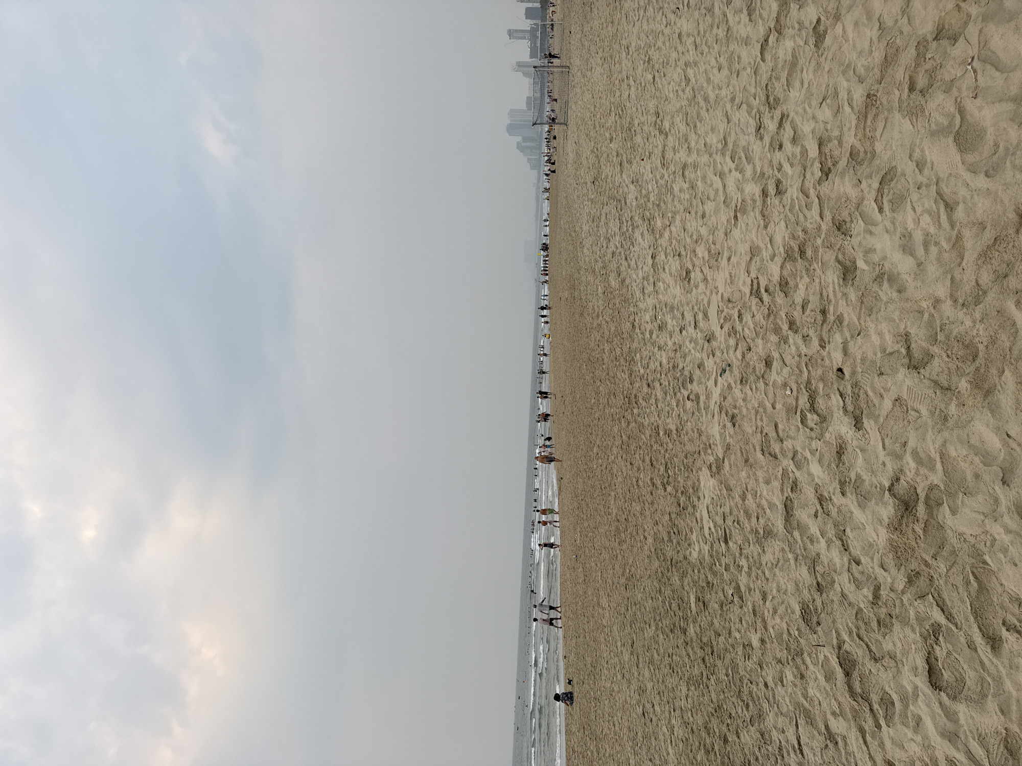 This is a wide-angle, outdoor photograph of a beach scene in Đà Nẵng, Vietnam. The sky is overcast with light clouds. The beach is sandy and crowded with people, some near the water's edge and others further up the beach. In the distance, there are city buildings visible along the horizon. The focus is sharp across the entire frame, indicating a deep depth of field. Footprints are visible in the sand. There is a soccer goal on the beach.