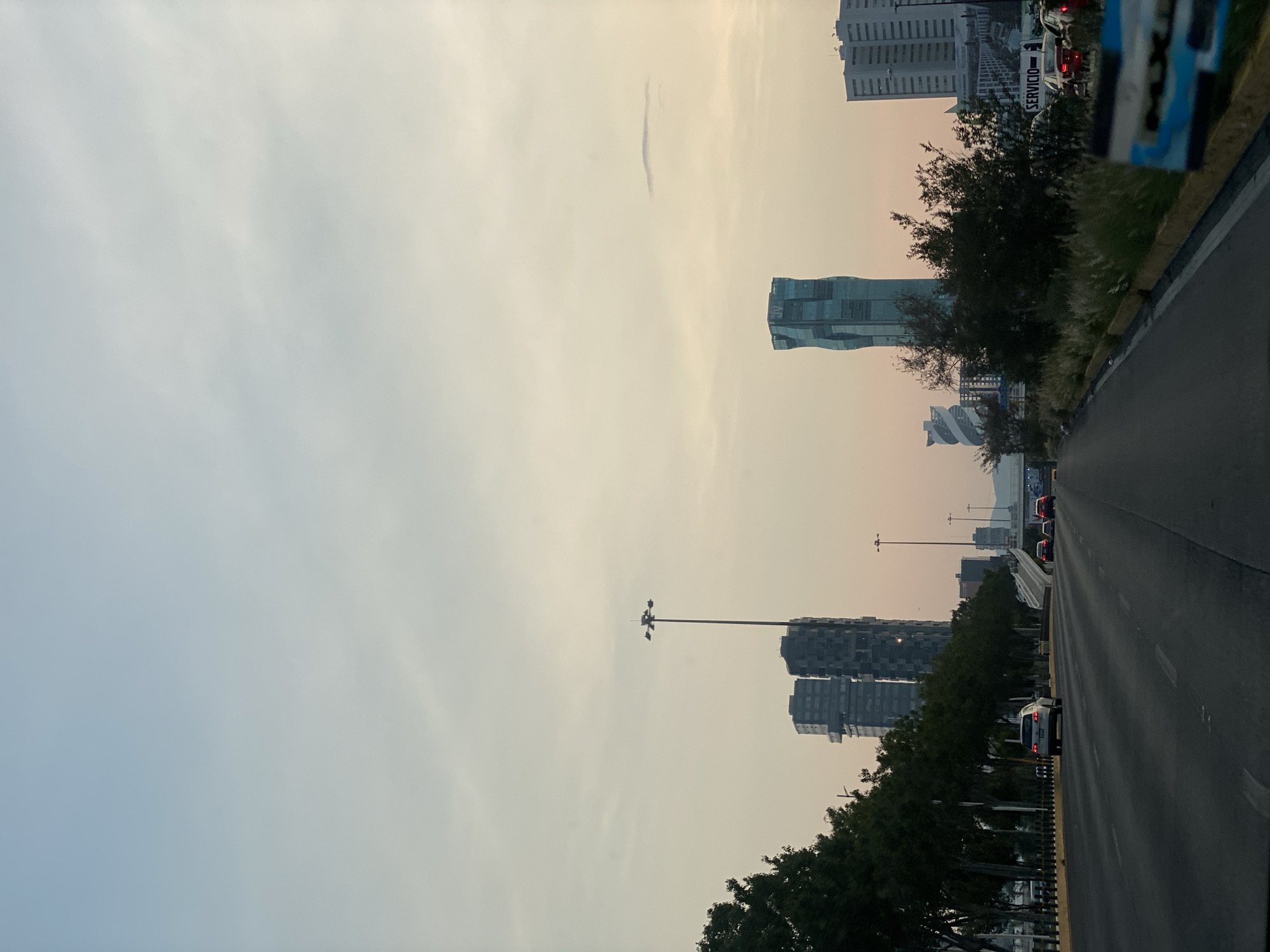 The image depicts a multi-lane road in an urban environment, with several tall buildings visible in the background. The sky is overcast with a light haze. Trees line the side of the road. Cars are present on the road. Streetlights are visible. The location is Unknown, Unknown.