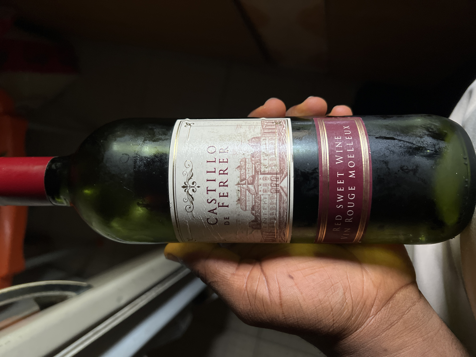 A hand is holding a green glass bottle of wine. The bottle has a label with text and an image of a building. The text on the label reads "CASTILLO DE FERRER" and below that "RED SWEET WINE VIN ROUGE MOELLEUX". The background is dark and out of focus, suggesting an indoor setting. The image does not contain any information about its location in Lagos, Nigeria.