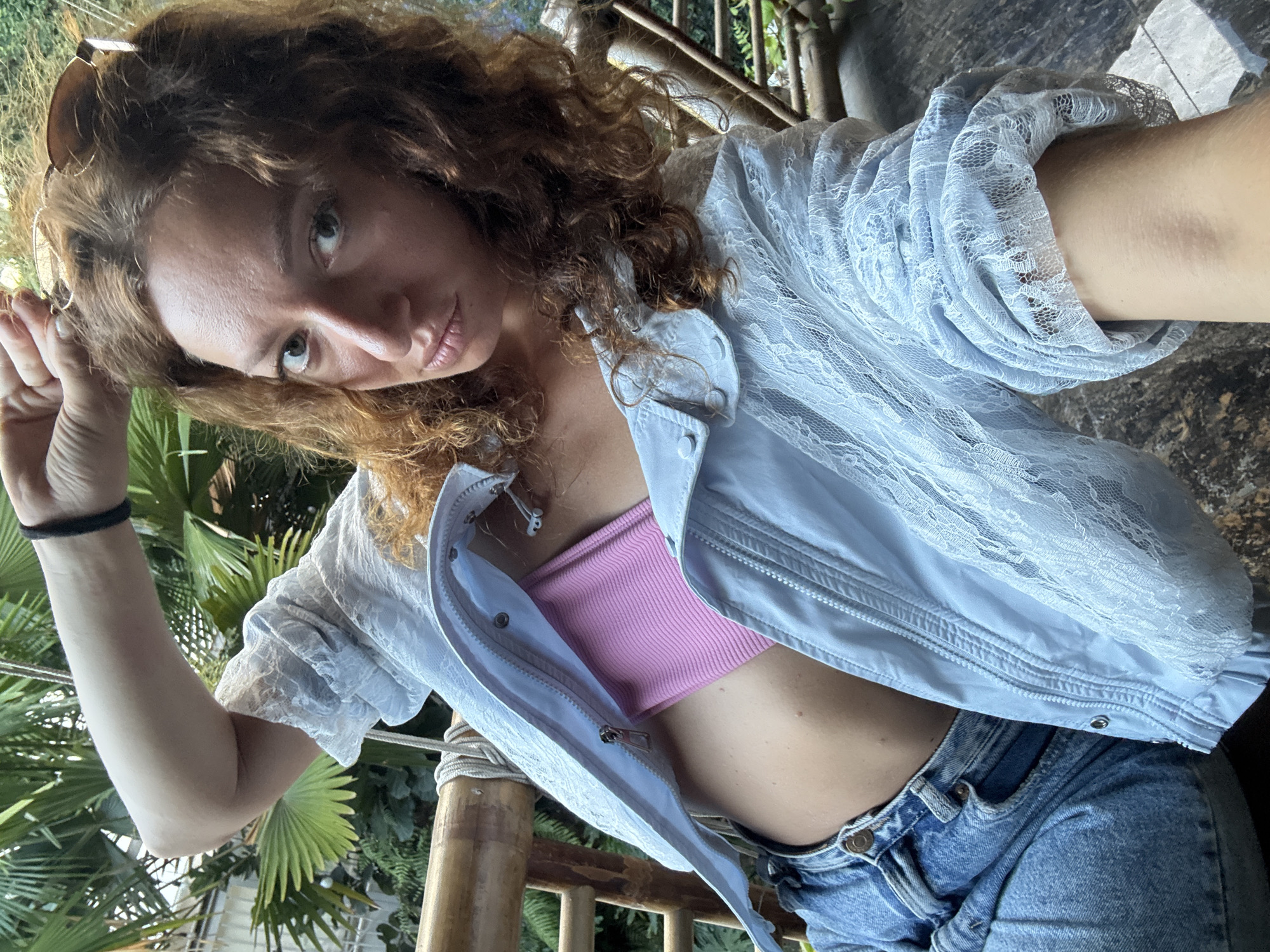 A medium shot captures a young adult female with curly reddish-brown hair and light eyes, wearing sunglasses pushed back onto her head, directly facing the camera. She is dressed in an unzipped light blue jacket featuring a lace overlay, revealing a pink ribbed bandeau top underneath. Her lower attire consists of light blue denim jeans. Her left arm is raised with a bent elbow, revealing a black hair tie on her wrist. The background consists of dense green tropical foliage, including large palm fronds, and visible bamboo-like wooden railing or furniture with ropes. The scene is set in an outdoor or semi-outdoor environment, consistent with a tropical climate, located in Trung Tam Ward, Vietnam.
