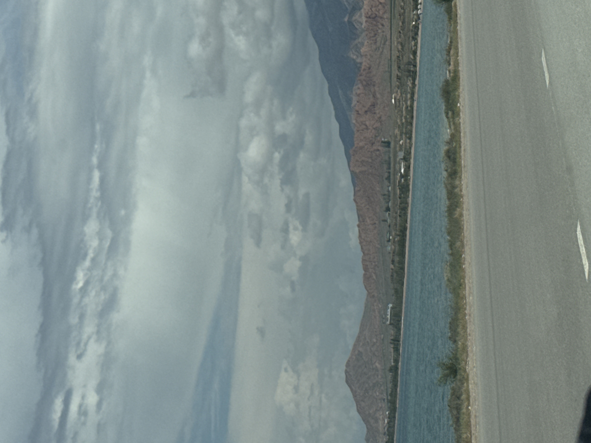 The image depicts a scenic view of Lake Issyk-Kul with mountains in the background. The foreground features a paved road with white lane markings, suggesting it is a highway or a main road. The road surface appears to be asphalt. Beyond the road, there is a grassy verge with some sparse vegetation.
The middle ground is dominated by the body of water, which is a vibrant turquoise color, characteristic of a lake. The water appears calm with some gentle ripples. Along the shoreline of the lake, there is a line of green trees and shrubs, indicating a vegetated area. Further inland, the landscape rises into hills and mountains. These mountains are a mixture of reddish-brown and darker, bluish-grey hues, with visible textures suggesting eroded rock formations. Some low-lying buildings or structures are scattered along the base of the mountains, possibly representing a settlement or resort area in the vicinity of Tong, Kyrgyzstan.
The sky is overcast with a layer of grey and white clouds, creating a diffused light that softens the overall scene. The clouds are varied in texture, with some appearing wispy and others more dense.
The image appears to have been taken from a moving vehicle, as suggested by the slight blur and the perspective of the road in the foreground. The overall atmosphere is one of natural beauty and tranquility, typical of a lakeside mountain landscape.