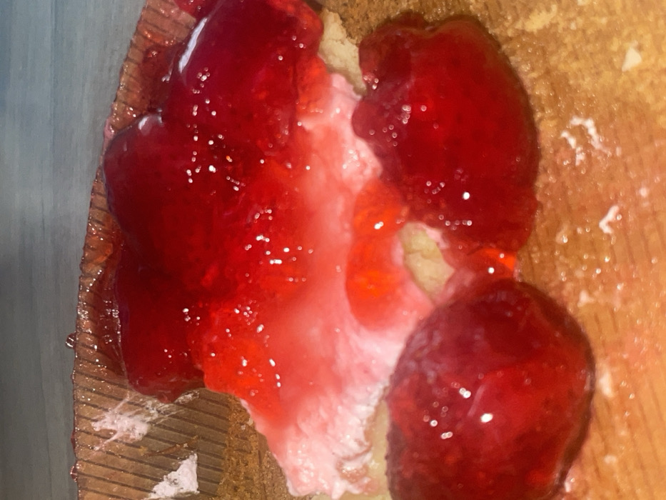A close-up shot shows a dessert item, likely a tart or pastry, topped with a generous layer of red, glistening jelly or fruit preserve. The base appears to be a light-colored pastry or cream, partially obscured by the topping. The surface beneath the dessert shows a wooden cutting board or tray. Some stray, translucent fibers are present on the board's edge, possibly related to the dessert itself. The overall image has a slightly tilted perspective, emphasizing the texture and detail of the sweet treat.
