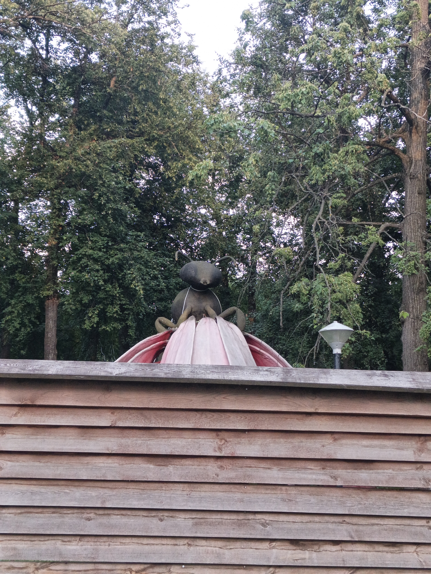 A statue resembling a large, dark insect, possibly a beetle, is perched atop a pink and red petal-like structure. The insect has large, bulbous eyes and what appear to be thin antennae. It is positioned in front of a dense background of green trees, with a few branches extending into the frame. A wooden fence, constructed from horizontal planks, runs across the bottom of the image, partially obscuring the base of the statue. A modern-looking outdoor lamp with a white, conical shade is visible to the right of the insect statue. The scene appears to be outdoors, likely in a park or garden, given the presence of trees and the decorative statue. The lighting suggests it might be late afternoon or early evening, with a soft, diffused light. No people are visible in the image.