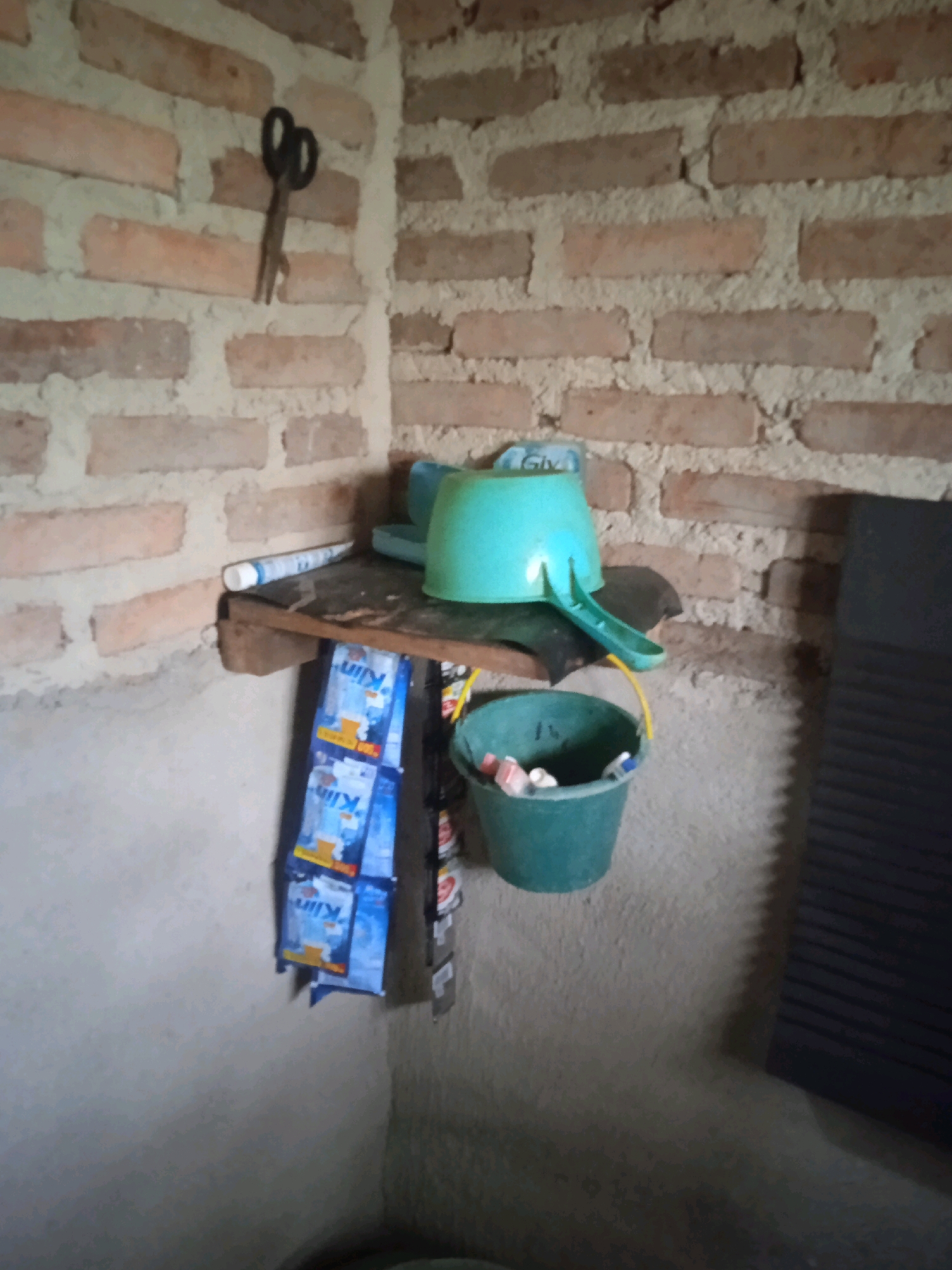 The image depicts a cluttered corner of a room with unplastered brick walls and a concrete floor. On the wall, a pair of black scissors hangs. Below the scissors, a wooden shelf is mounted, holding a tube of toothpaste, a teal-colored basin, and a smaller teal bucket. The bucket contains several small, colorful items. Hanging from the shelf are multiple blue and yellow packets of "Klin" powdered milk. To the right of the shelf, a dark, ribbed object, possibly a washboard, leans against the wall. The overall impression is of a utilitarian space, possibly a bathroom or kitchen in a rural or developing area of Karang Sari, Indonesia.