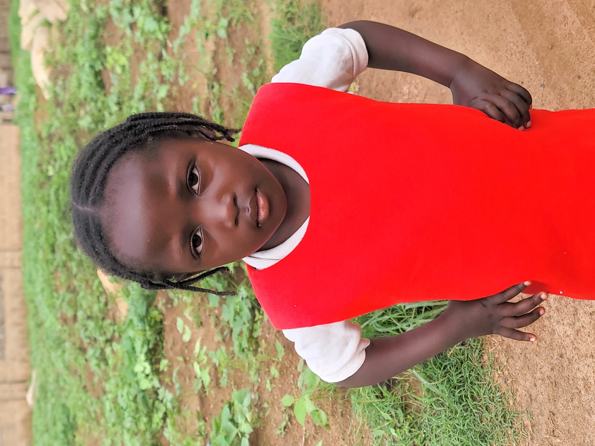 A young girl with braided dark hair stands outdoors in Barakin Akawo, Nigeria. She is wearing a white shirt under a bright red sleeveless dress. Her hands are placed on her hips, and she is looking directly at the camera with a neutral expression. The background consists of a field with green plants and brown soil, and a blurred structure in the distance.