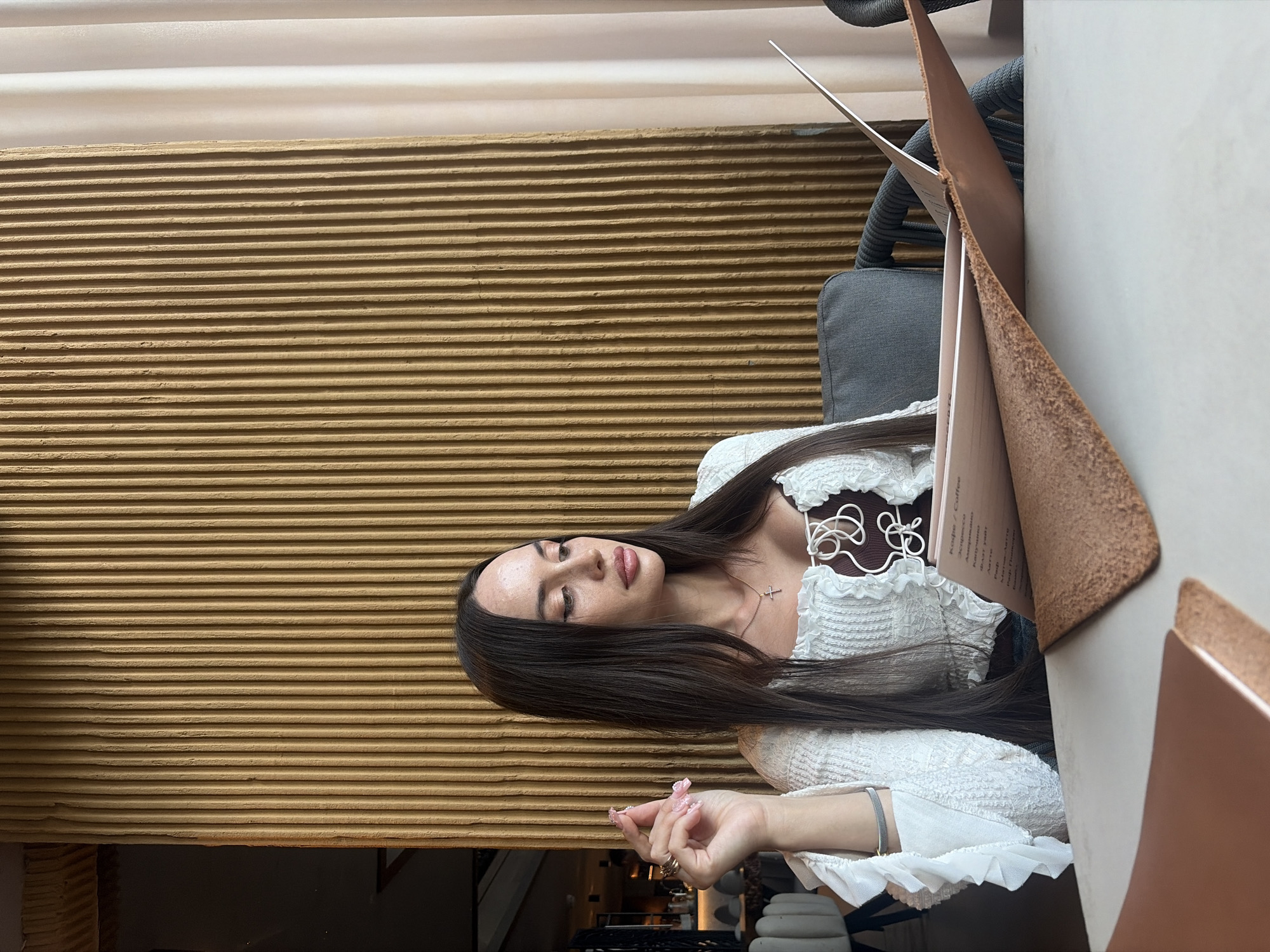 A woman is seated at a table in a restaurant, looking at a menu. She has long dark hair and is wearing a white ruffled top. The restaurant has a textured, ribbed wall in a warm, earthy tone behind her and pale curtains to the right. The table is white, and there are leather menu holders. The scene appears to be in Saint Petersburg, Russia, indicated by the menu text written in Cyrillic. The woman's hand is raised, showing painted fingernails and rings.
